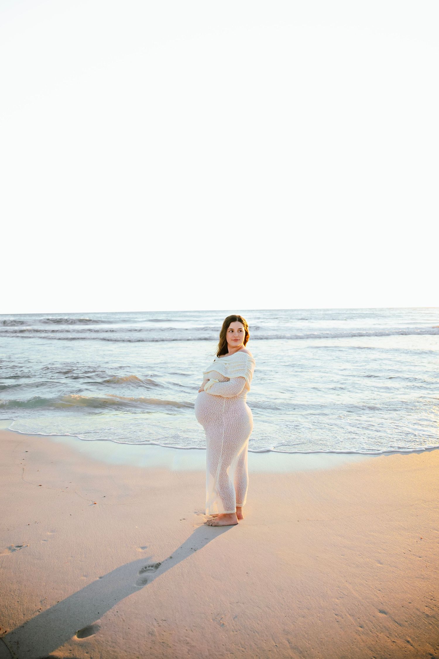 Pregnant woman in sheer white dress standing alone on beach at sunrise, cradling her baby bump.