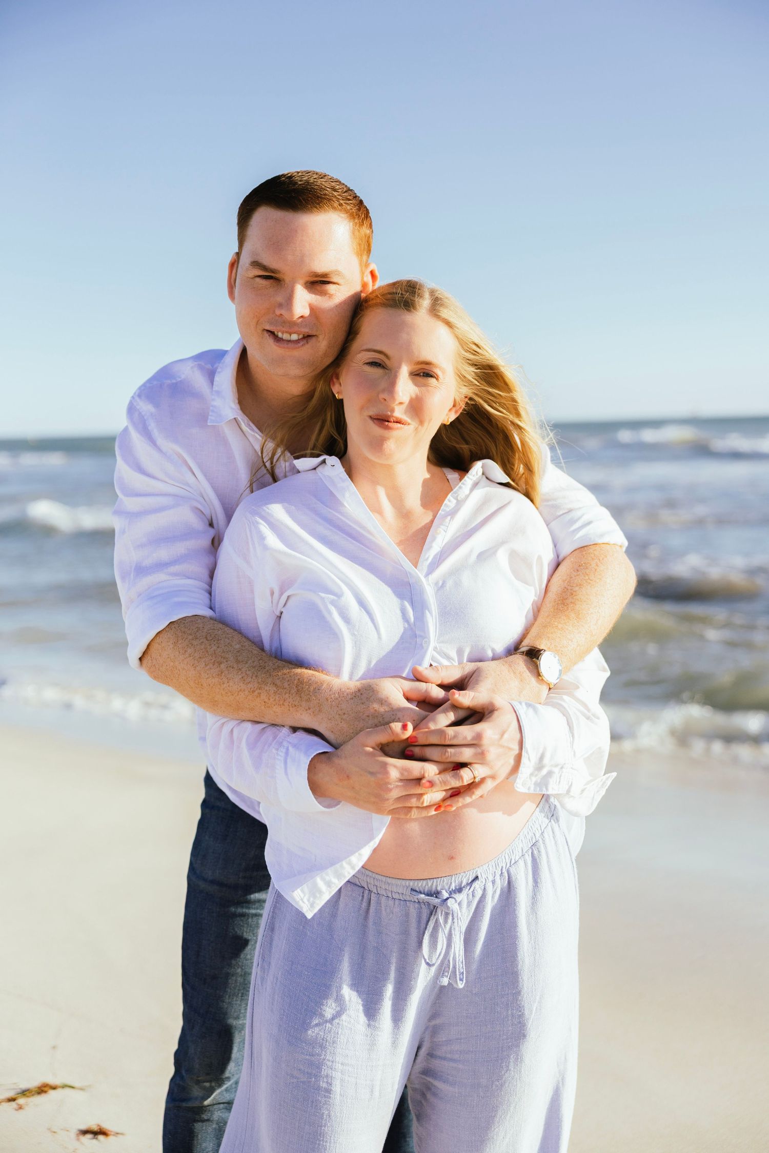 A happy couple embraces on a sunny beach, the woman visibly pregnant, both wearing white shirts with ocean waves behind them.