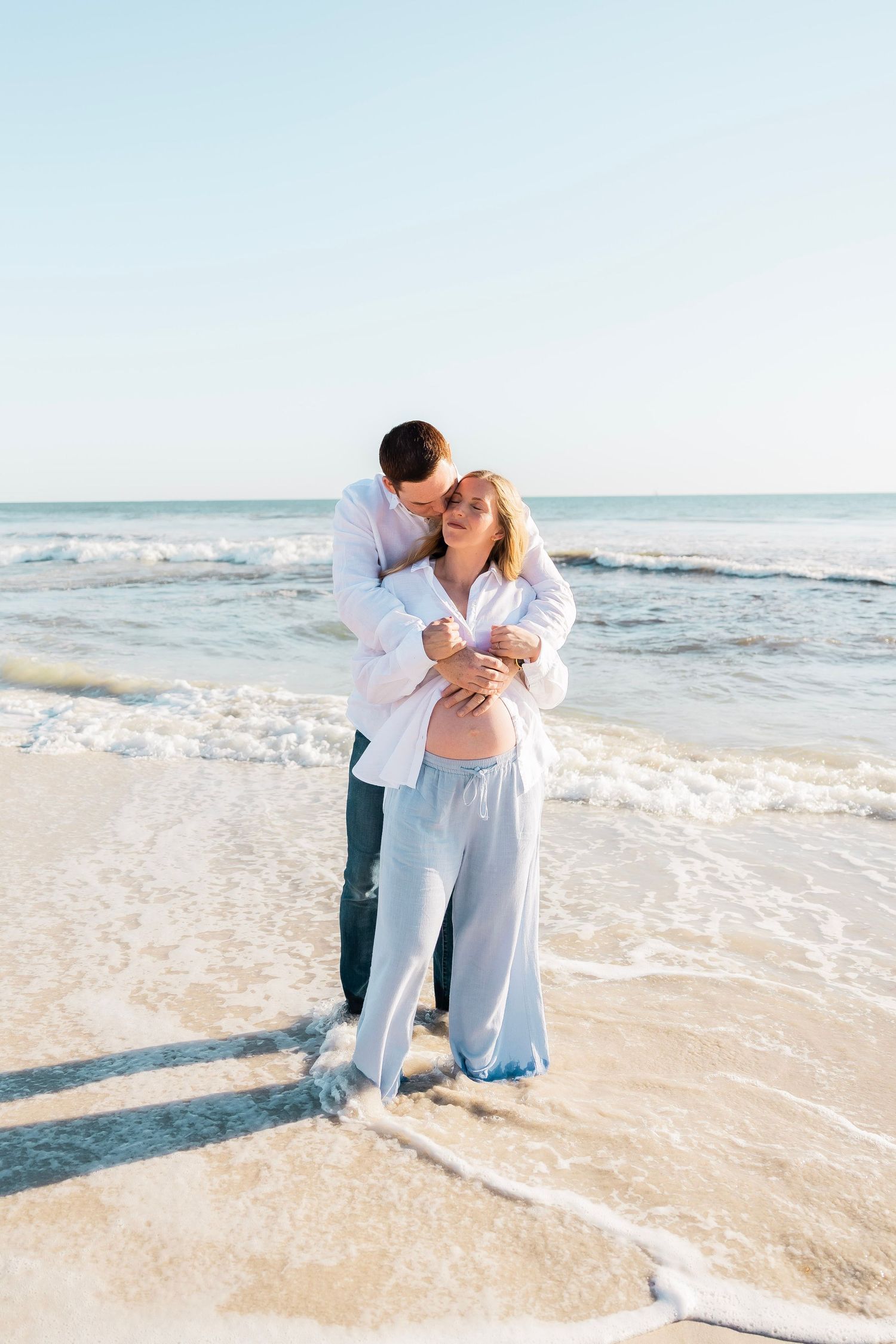 Couple sharing tender moment on beach, man kissing pregnant womans cheek by ocean waves.
