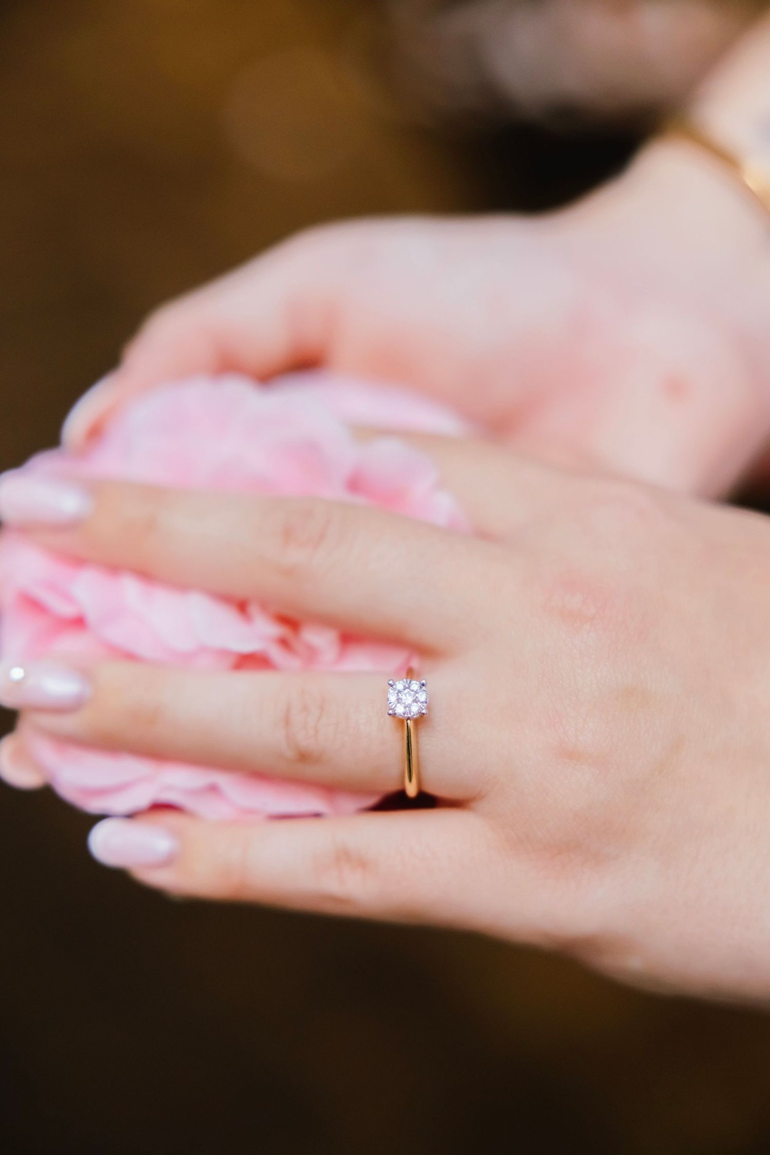 Close-up of a delicate gold solitaire diamond engagement ring on a hand holding pink petals.