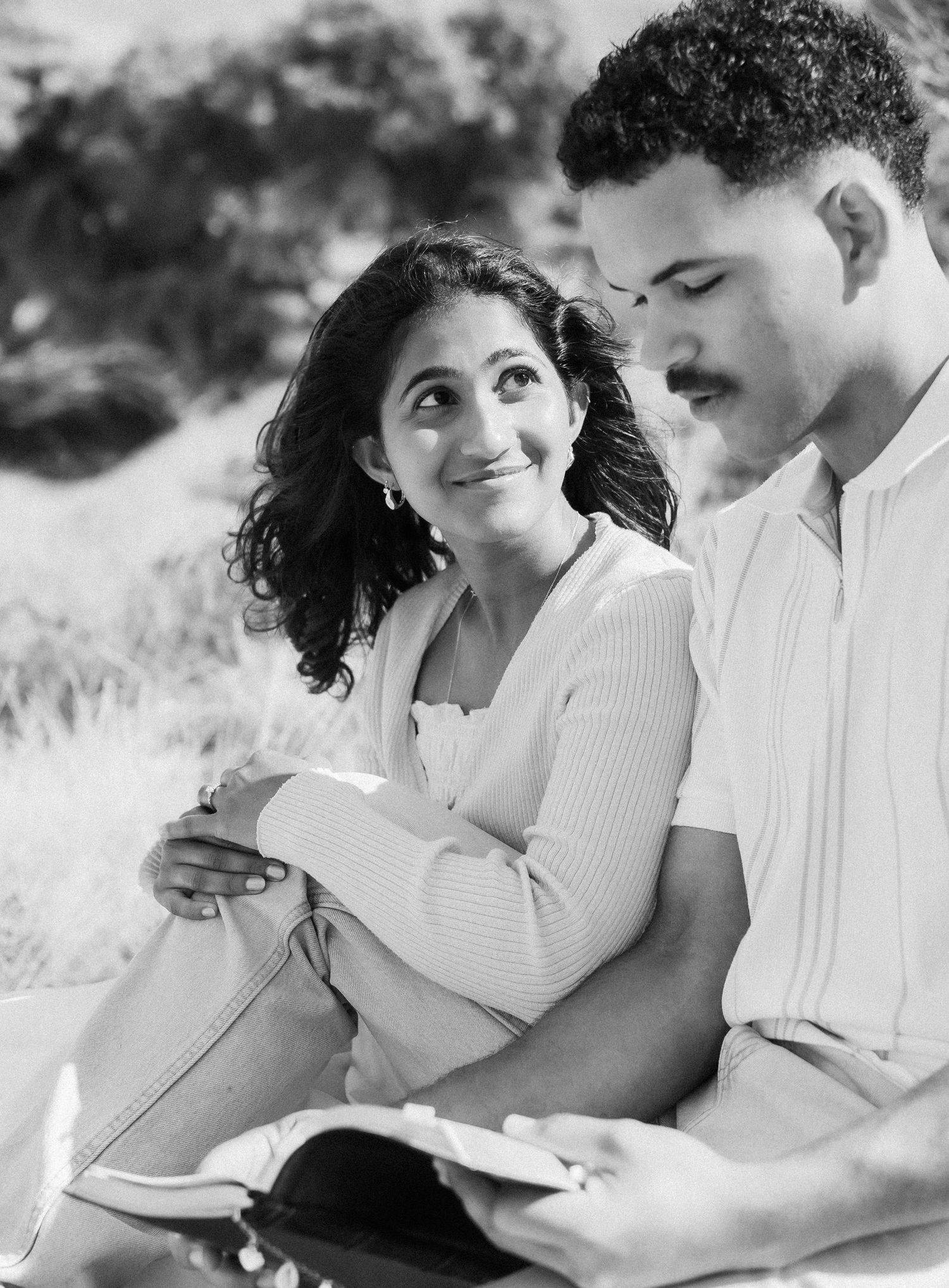 Black and white photo of a smiling woman leaning against a man while reading a Bible together outdoors.