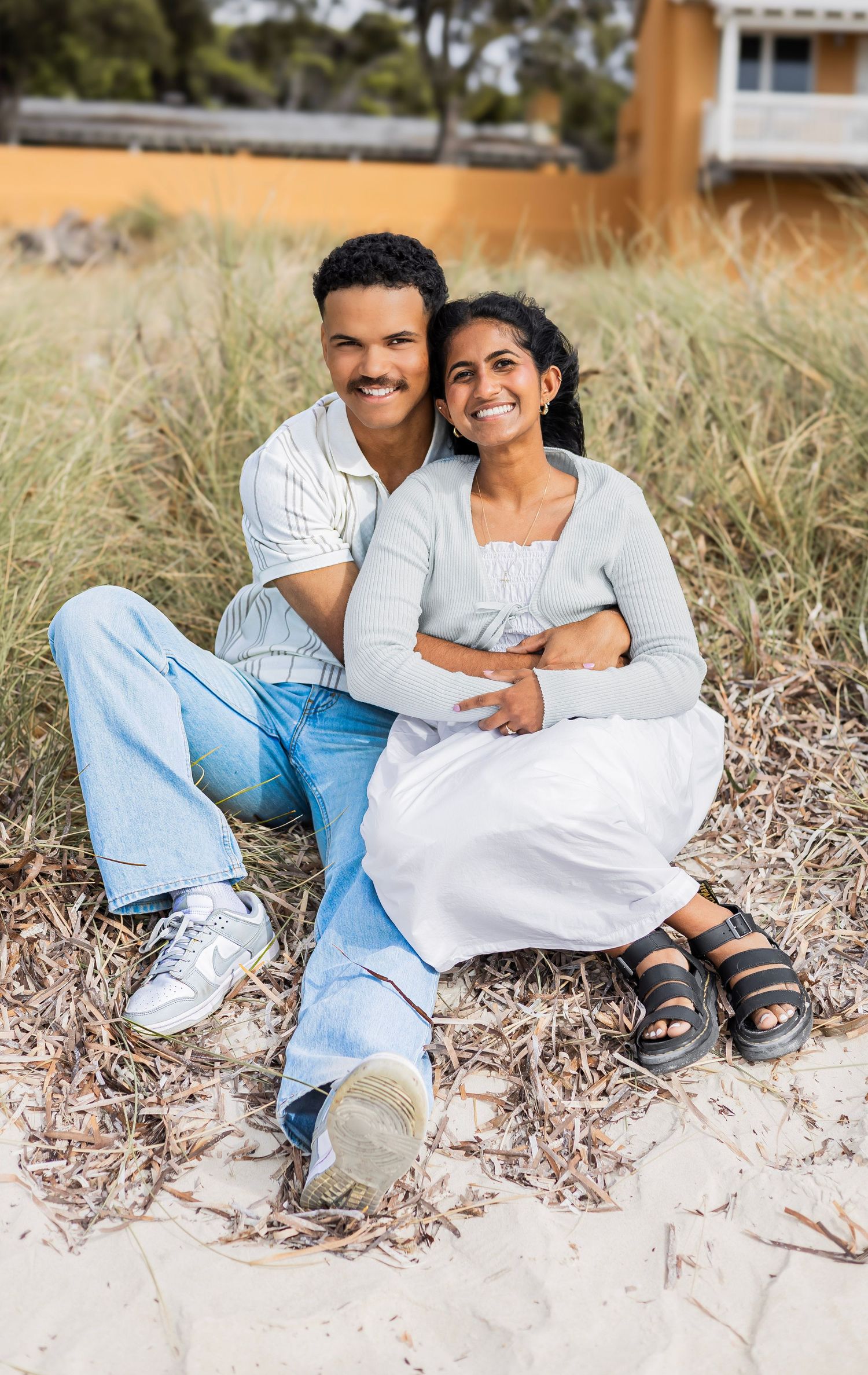 Happy couple sitting on beach sand dunes, man embracing woman from behind, both smiling warmly.