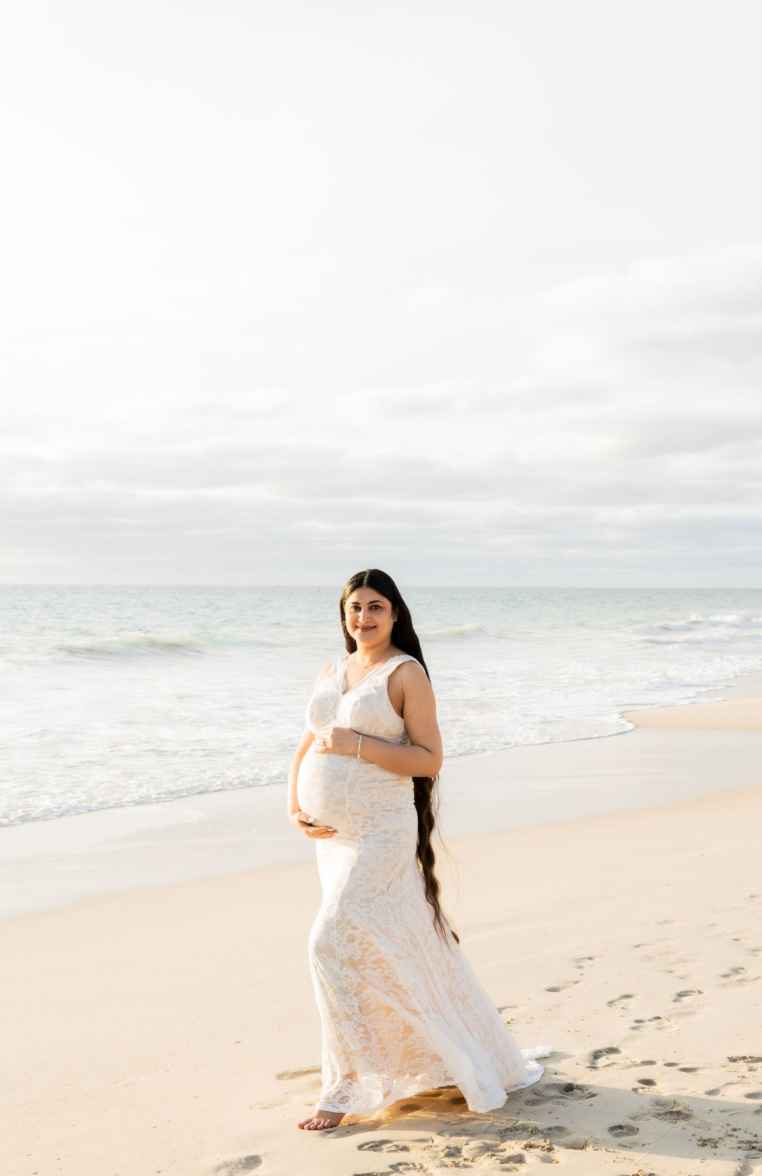 Pregnant woman in white lace gown cradling belly on a serene sandy beach.