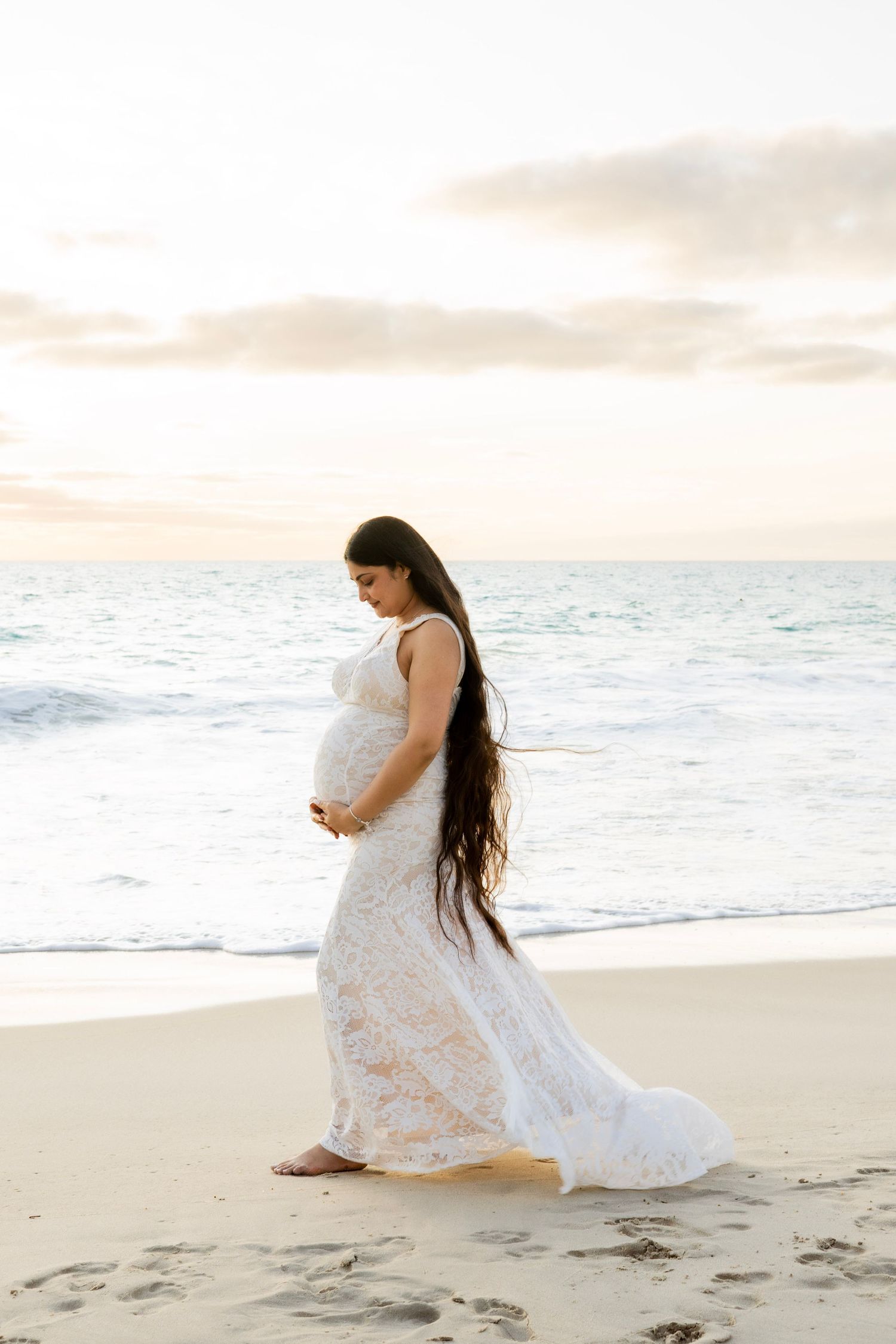 Pregnant woman in white lace dress walking on beach at sunset, holding her baby bump near ocean waves.
