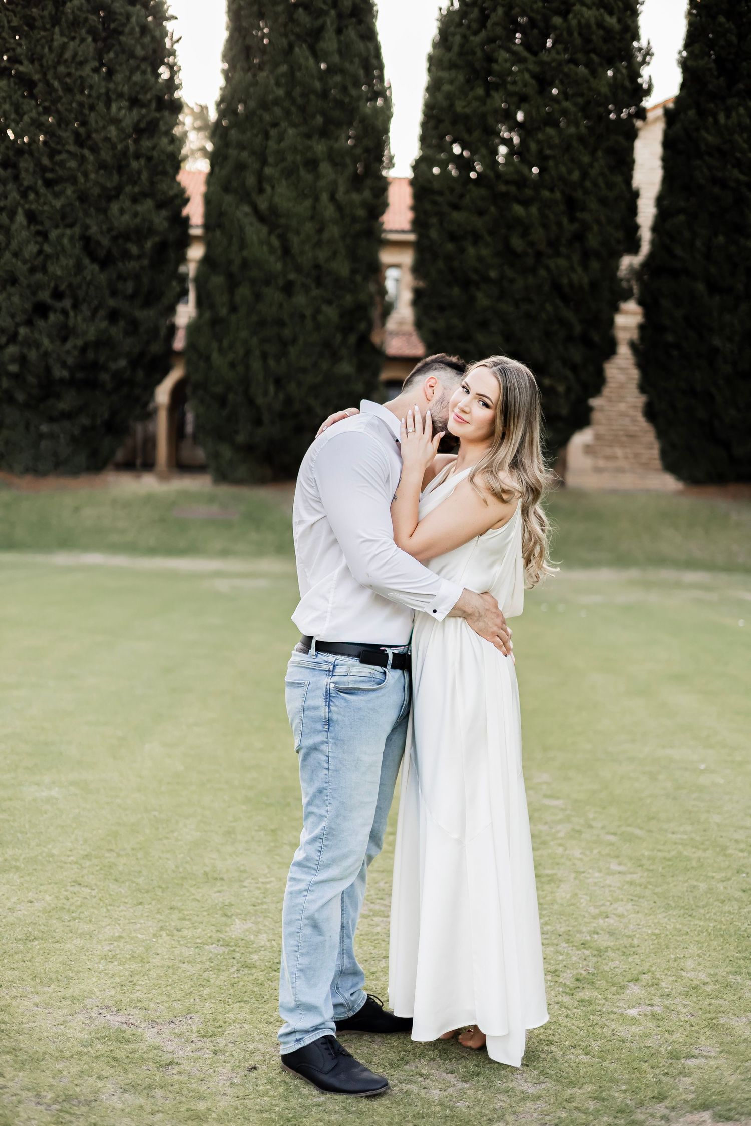 Couple embracing and smiling outdoors on lush green lawn with cypress trees and historic building.
