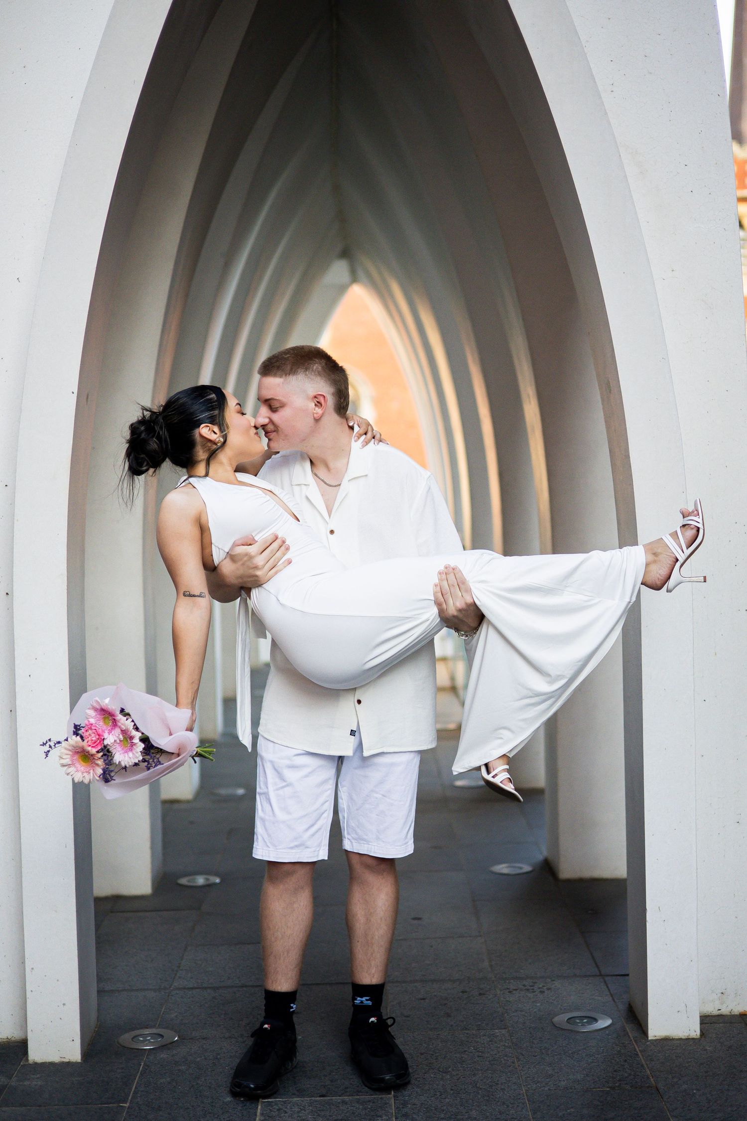 Groom lifting bride in white dress while kissing under white arches.