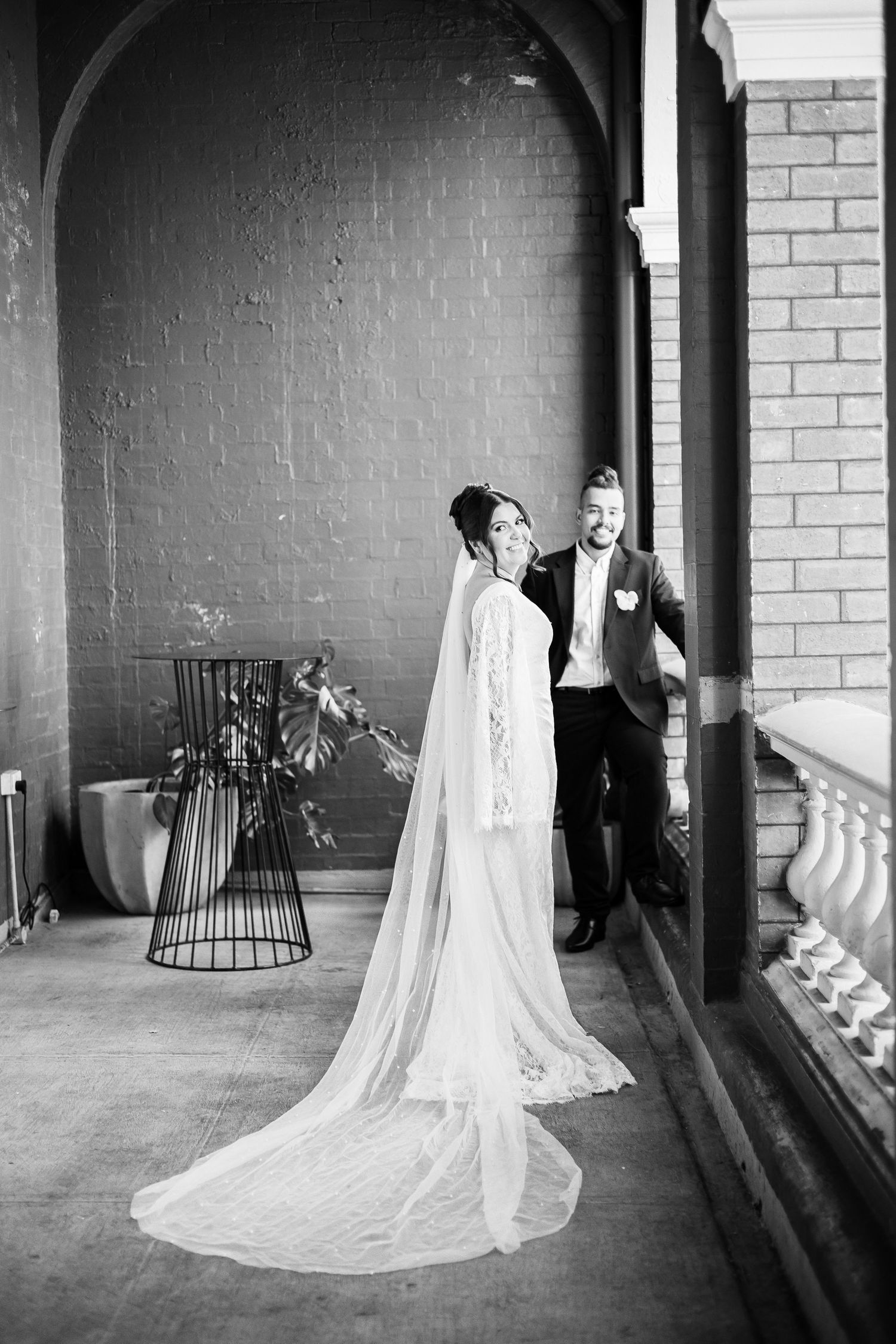 Black and white photo of bride with cathedral veil and groom standing on ornate brick building balcony.