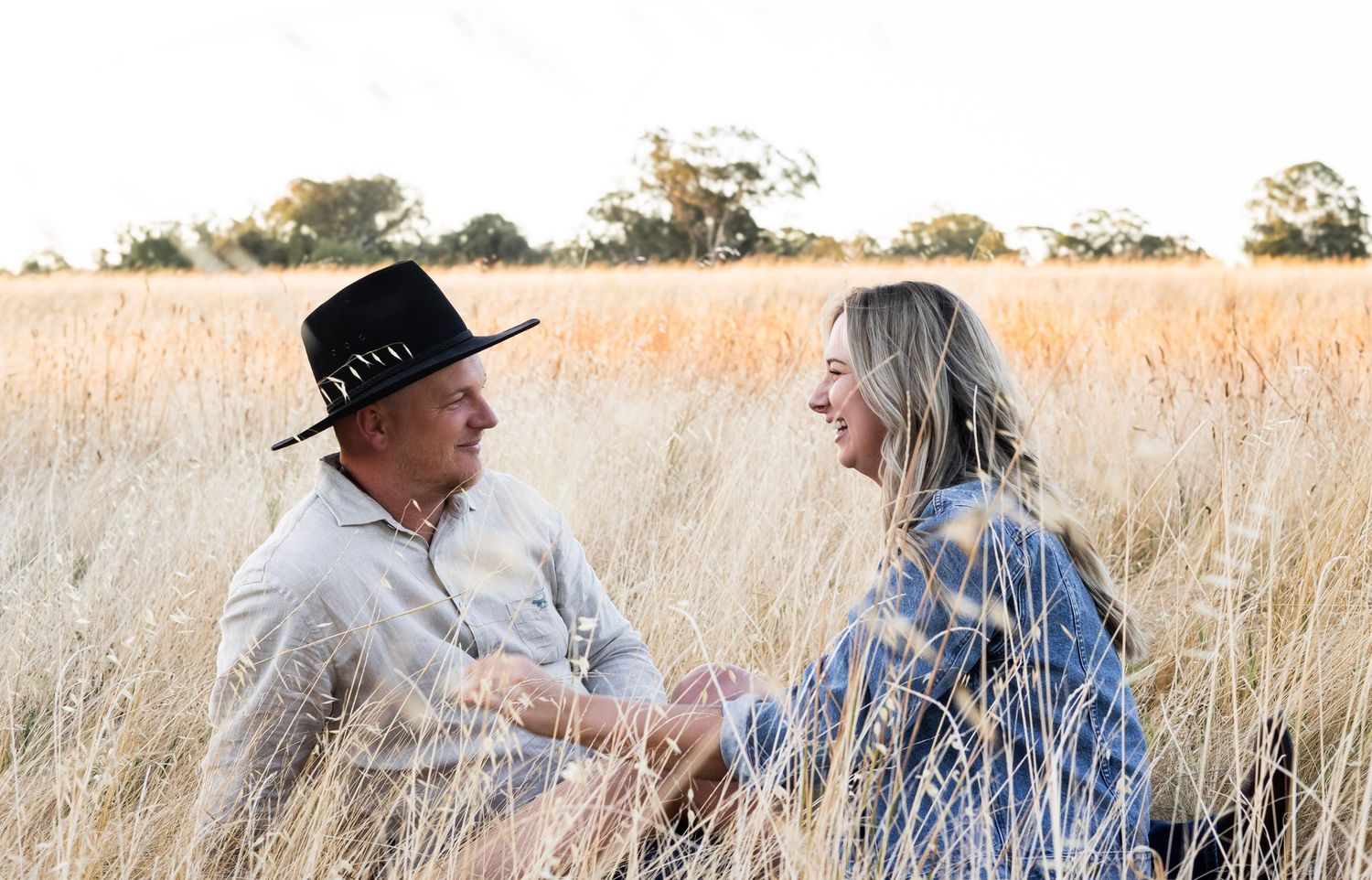 Couple sitting in tall golden grass, smiling and laughing together during a warm sunset session.