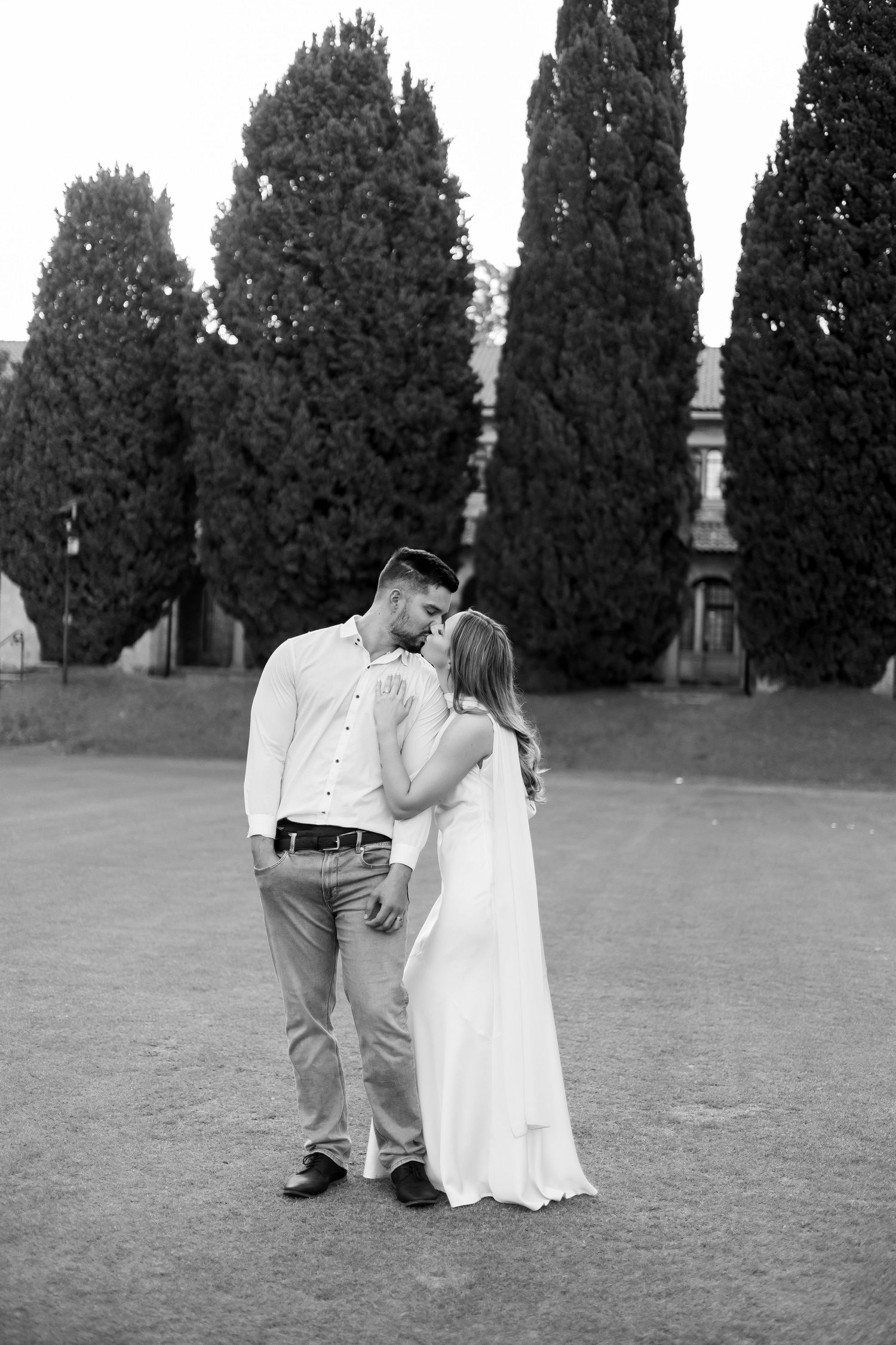 Black and white photo of couple sharing a kiss outdoors with tall cypress trees in background.