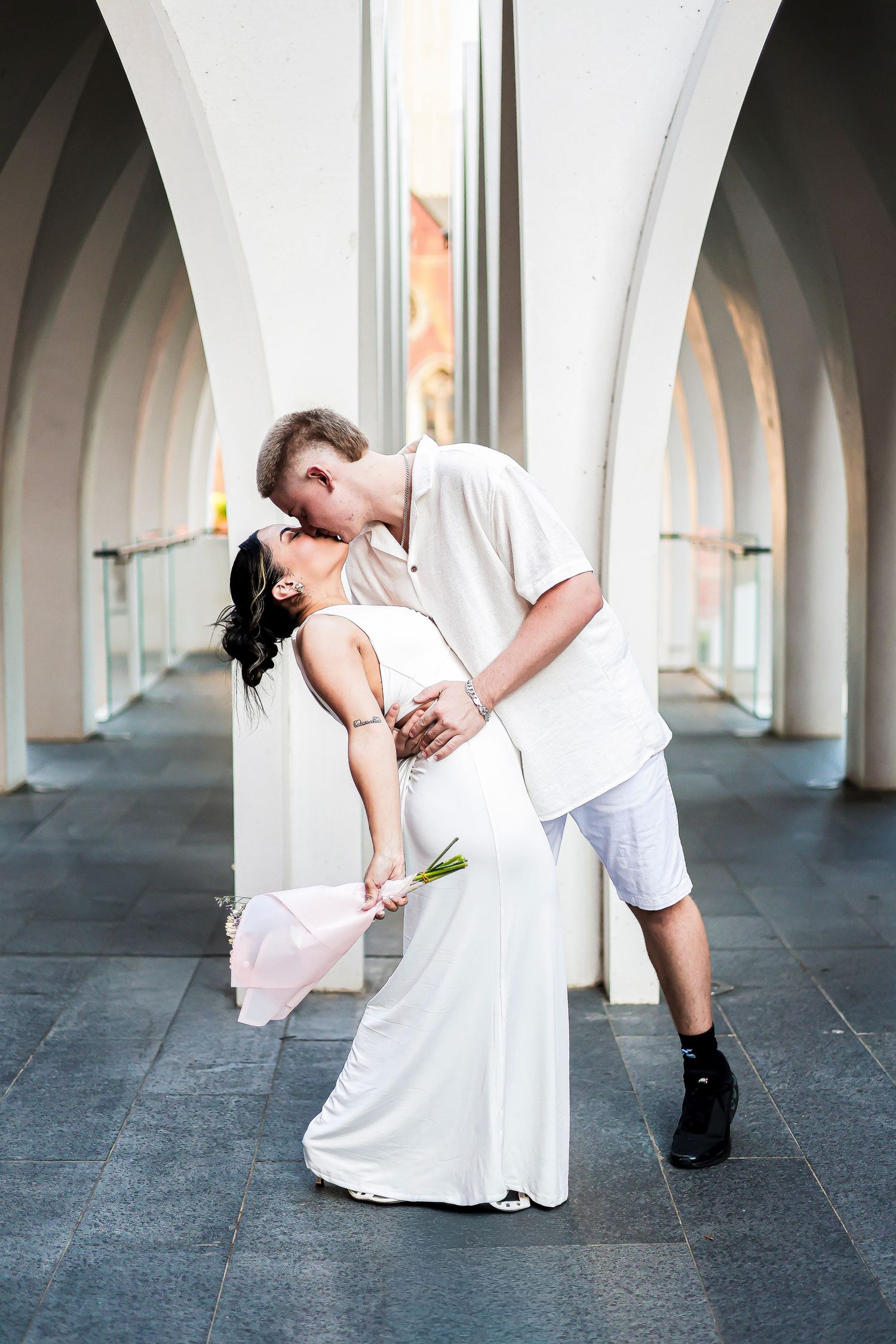 Couple sharing a romantic dip kiss in white wedding attire among elegant arched columns.