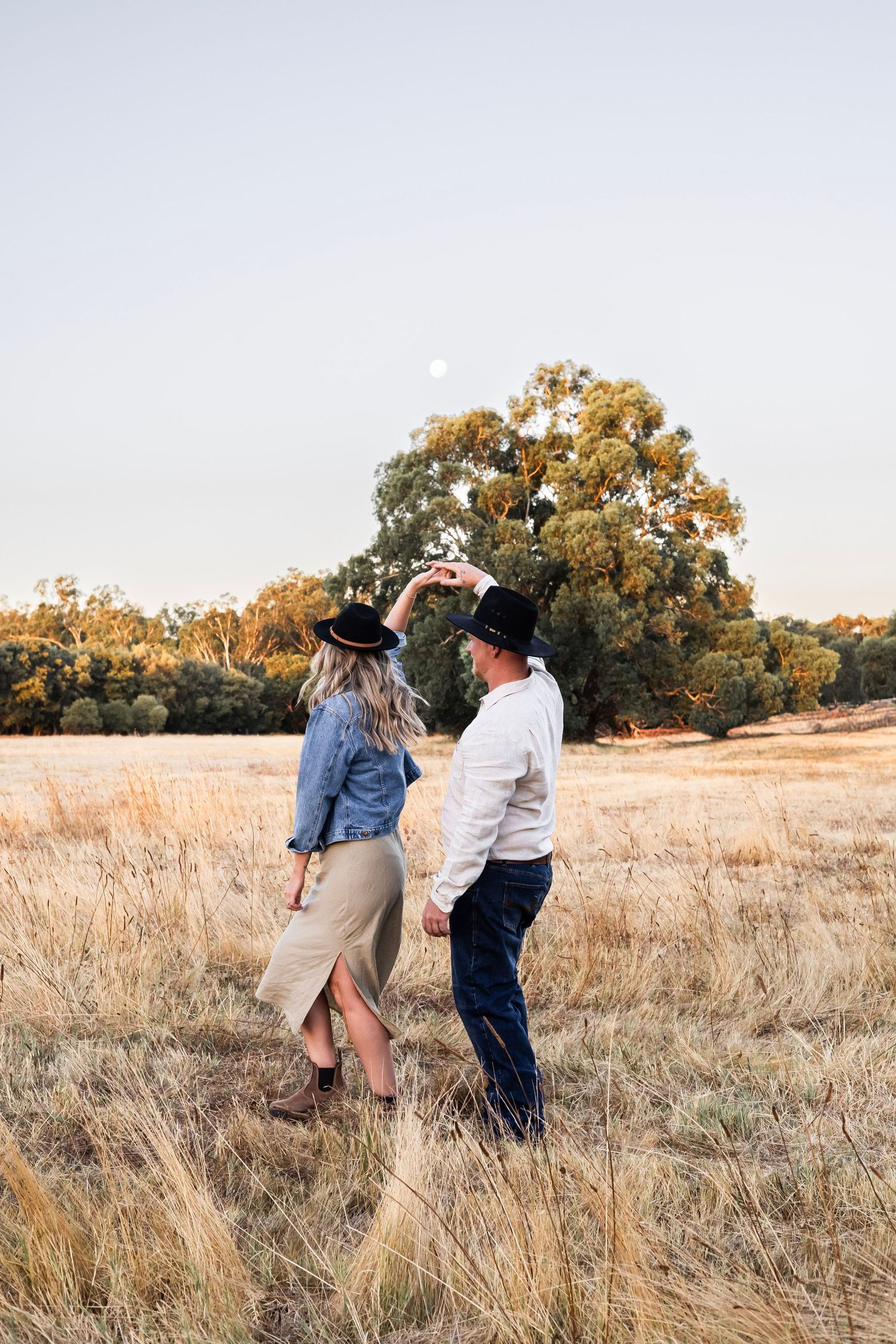 Couple dancing in a golden field at dusk, man spinning woman wearing cowboy hats under a full moon.