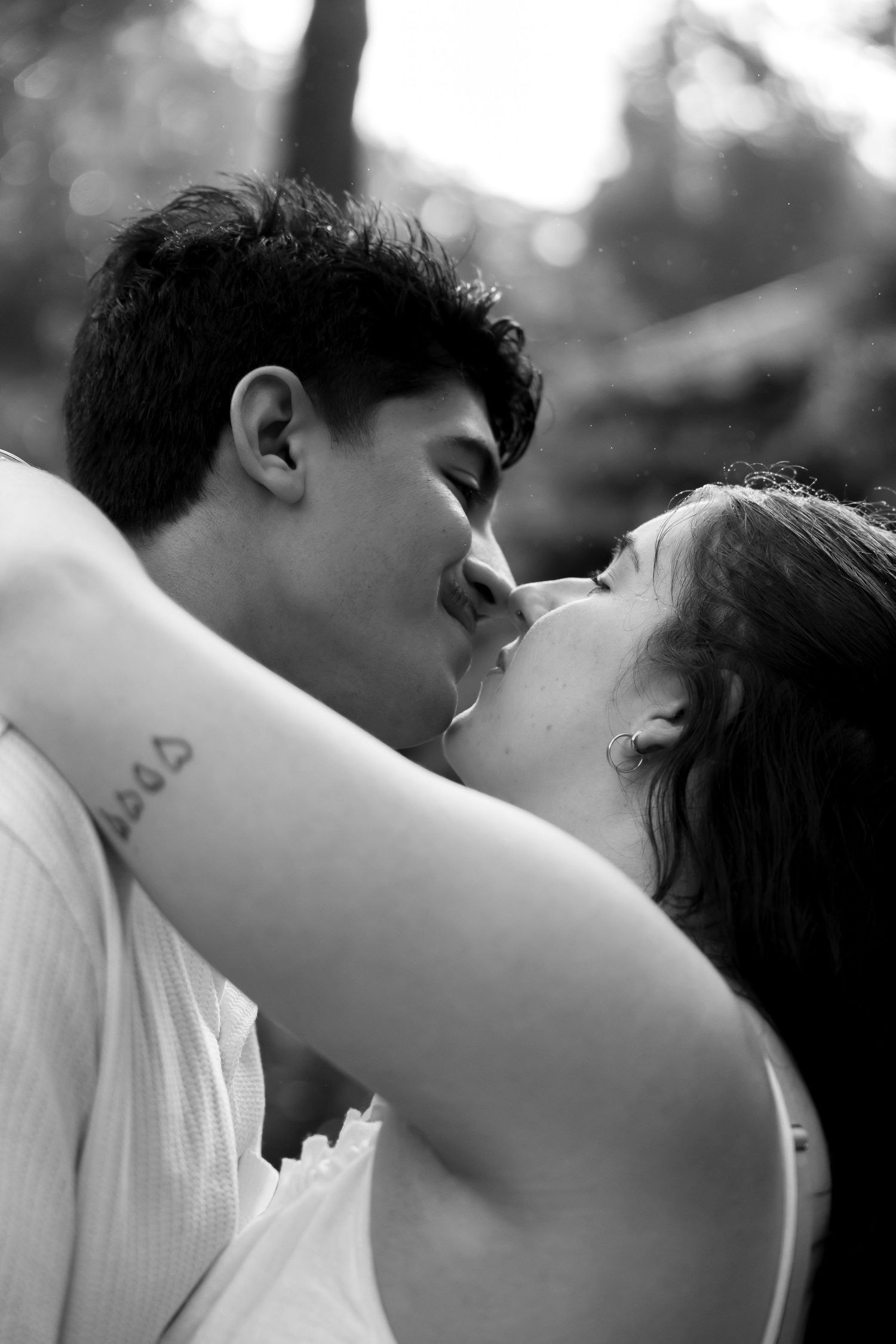 Black and white photo of couple kissing closely outdoors, woman with tattoo embracing mans neck.