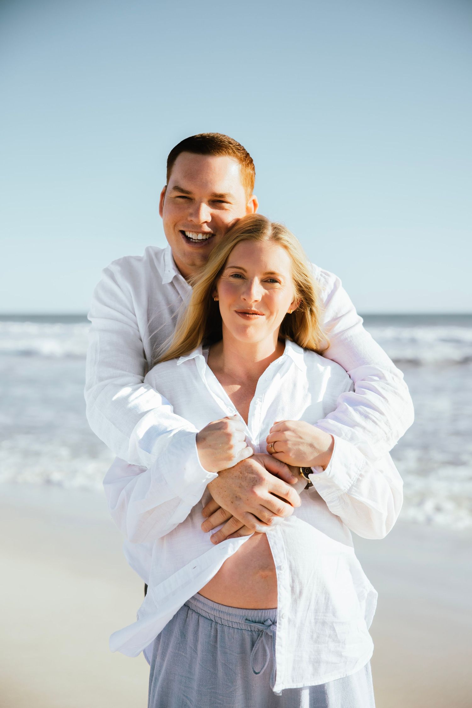 Couple in white shirts embracing on beach, pregnant woman showing baby bump near ocean waves.