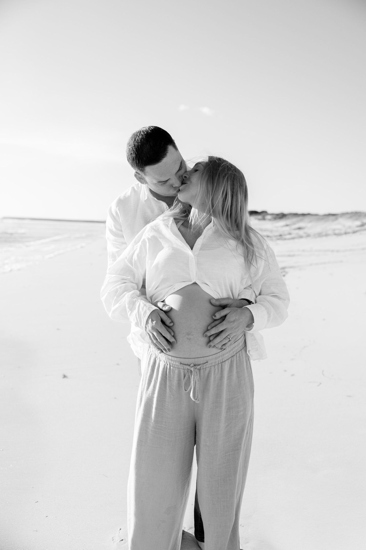 Black and white photo of a couple kissing on the beach, man embracing pregnant woman's belly.