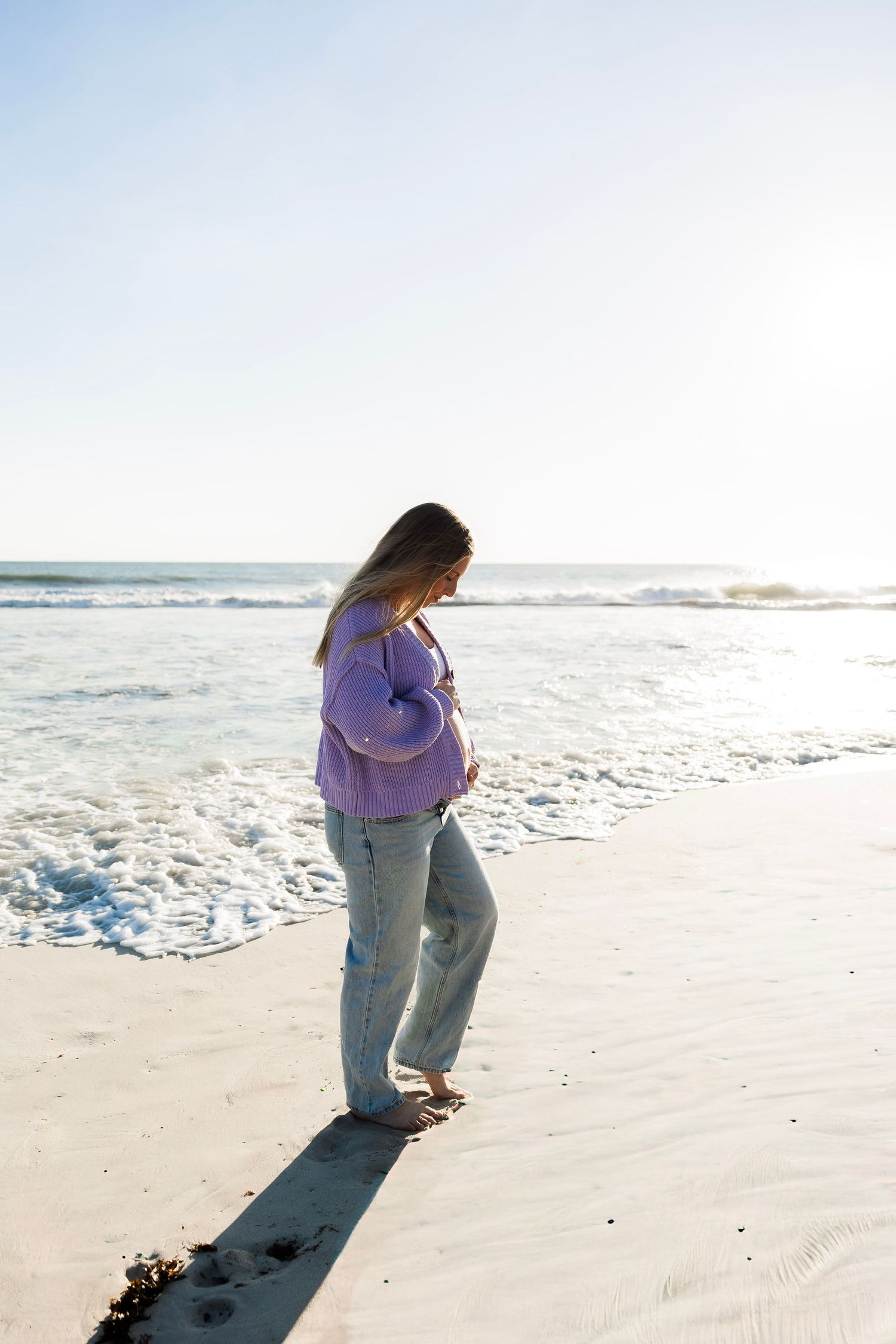 Pregnant woman in purple cardigan and jeans stands barefoot on sandy beach near ocean waves.