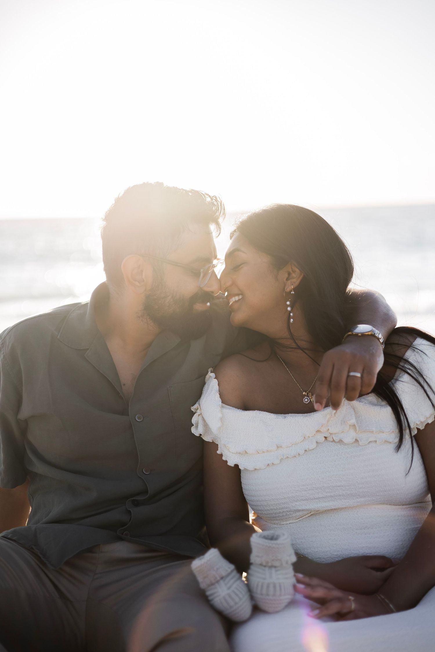 Pregnant couple sharing a tender moment on the beach at sunset, woman in white dress holding baby booties.