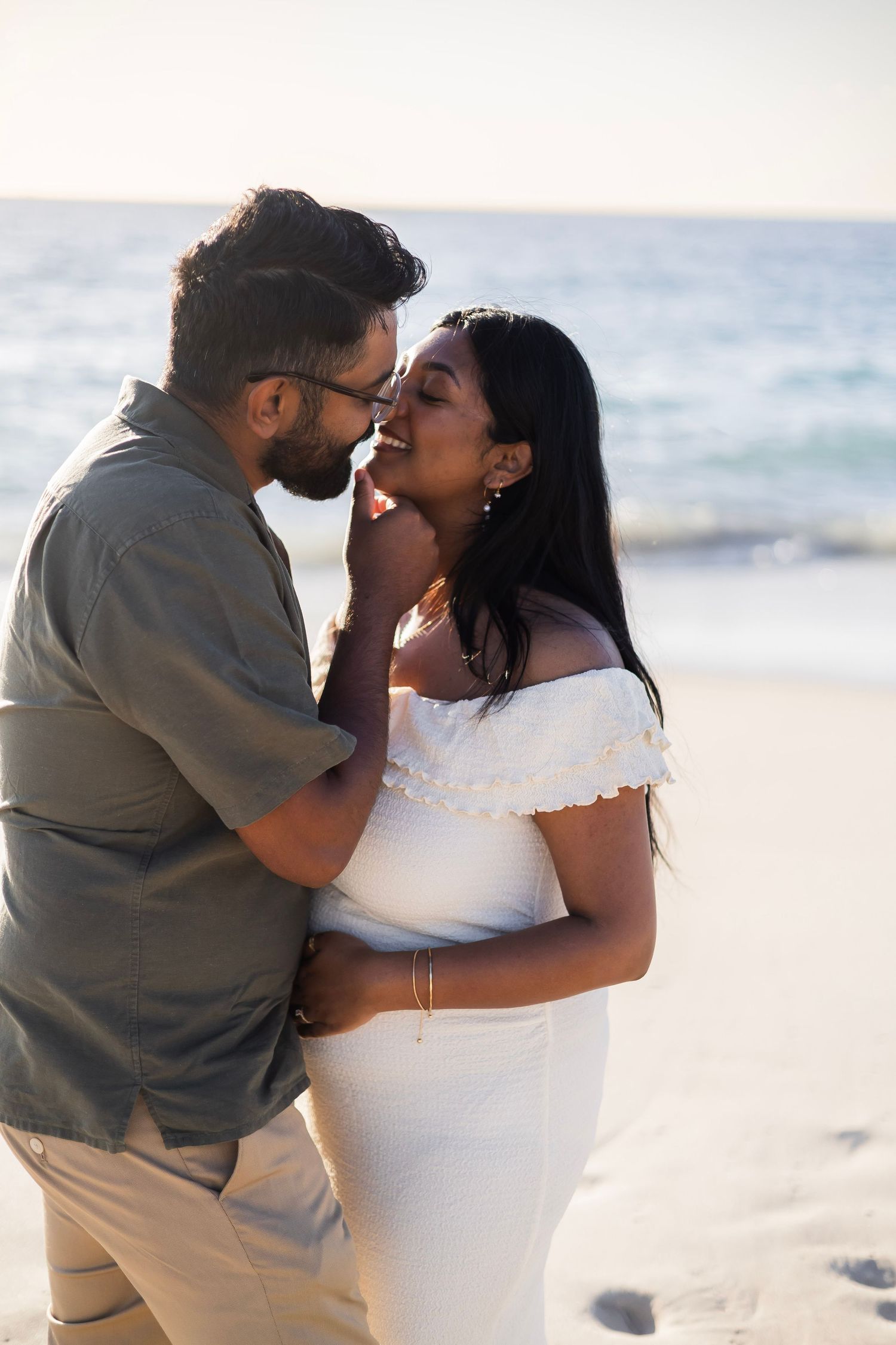 Couple sharing a romantic moment on the beach, man in gray shirt tenderly holding woman's face in white off-shoulder dress.