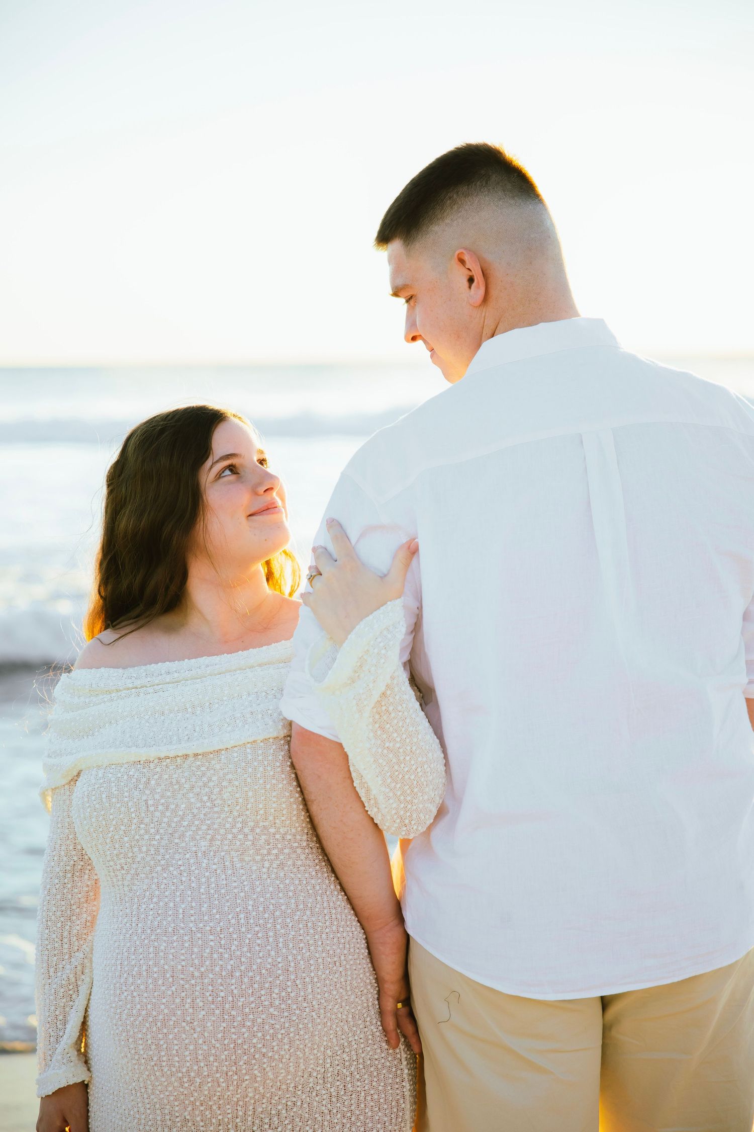 Pregnant couple embracing at beach during golden sunset, woman in white lace dress looking up at partner.