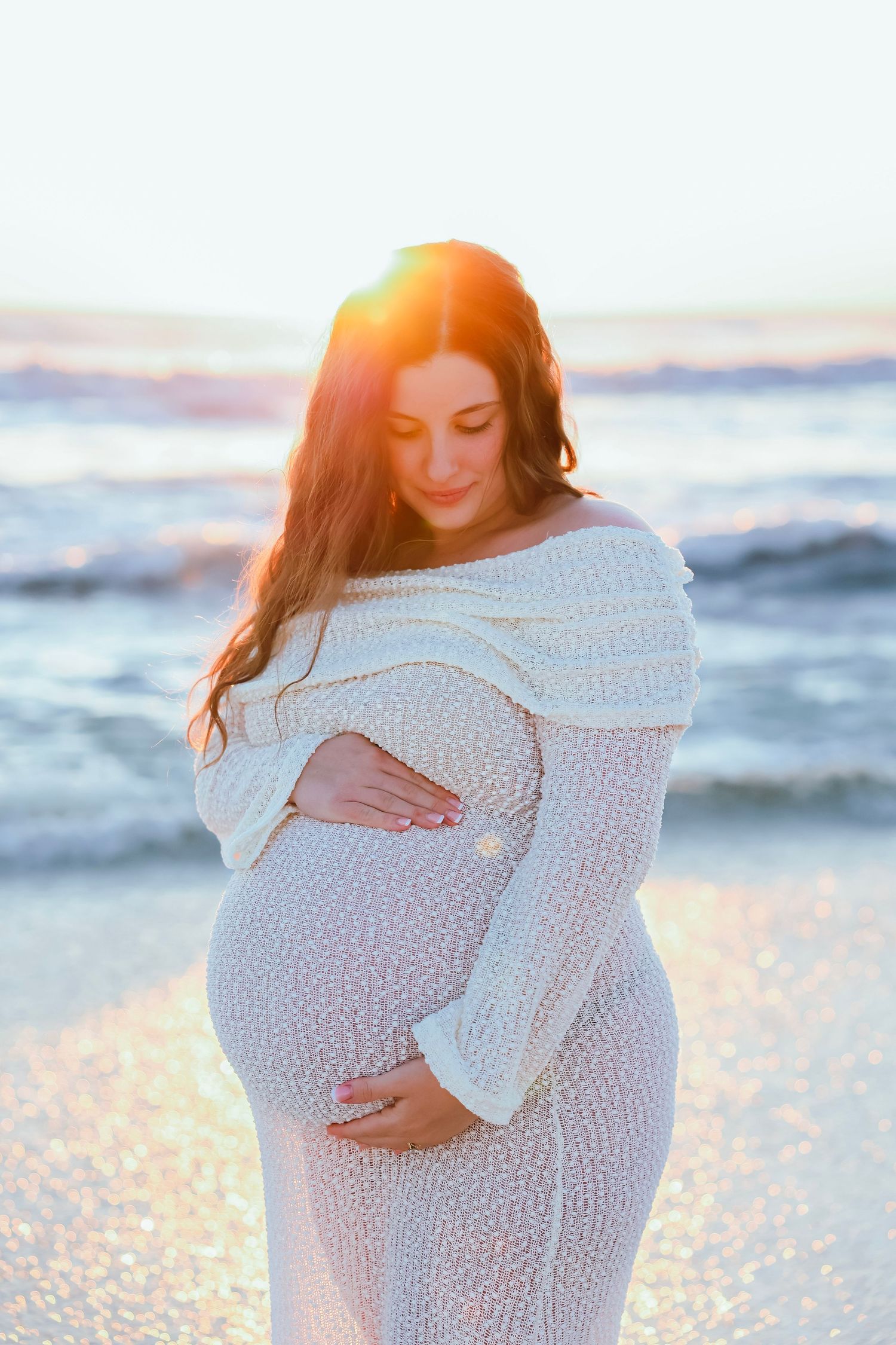 Pregnant woman in white lace dress cradling baby bump at golden hour beach sunset.