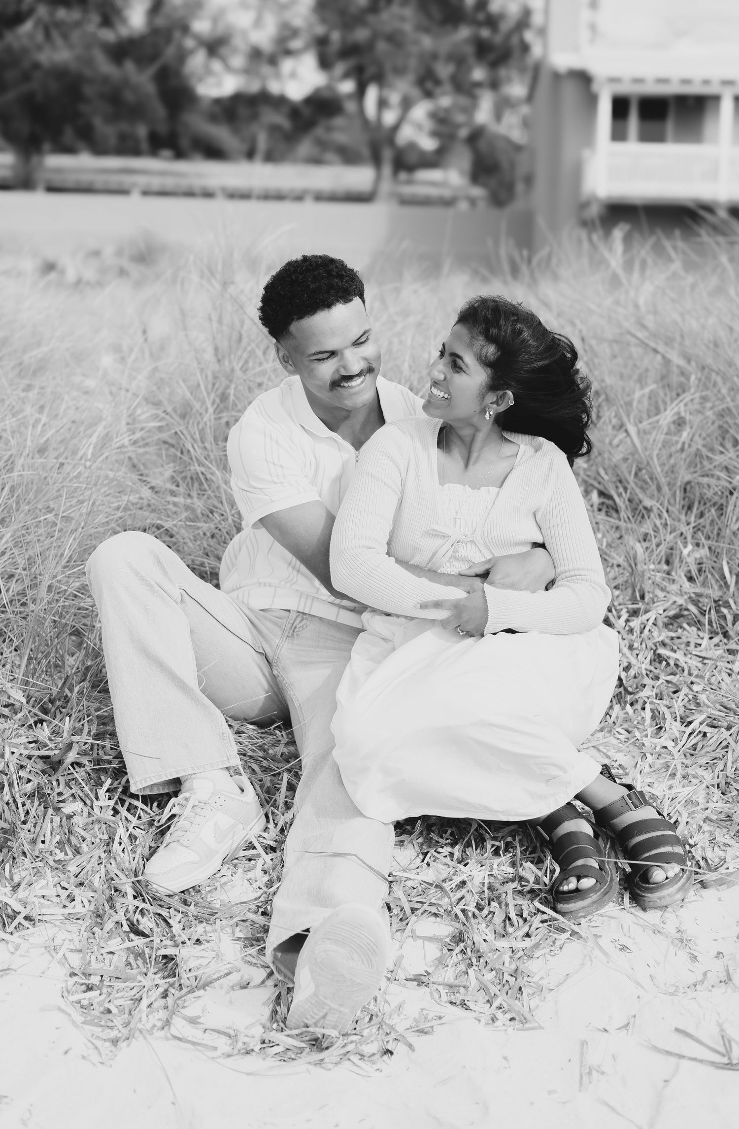 Black and white photo of romantic couple sitting in beach grass, gazing lovingly at each other.