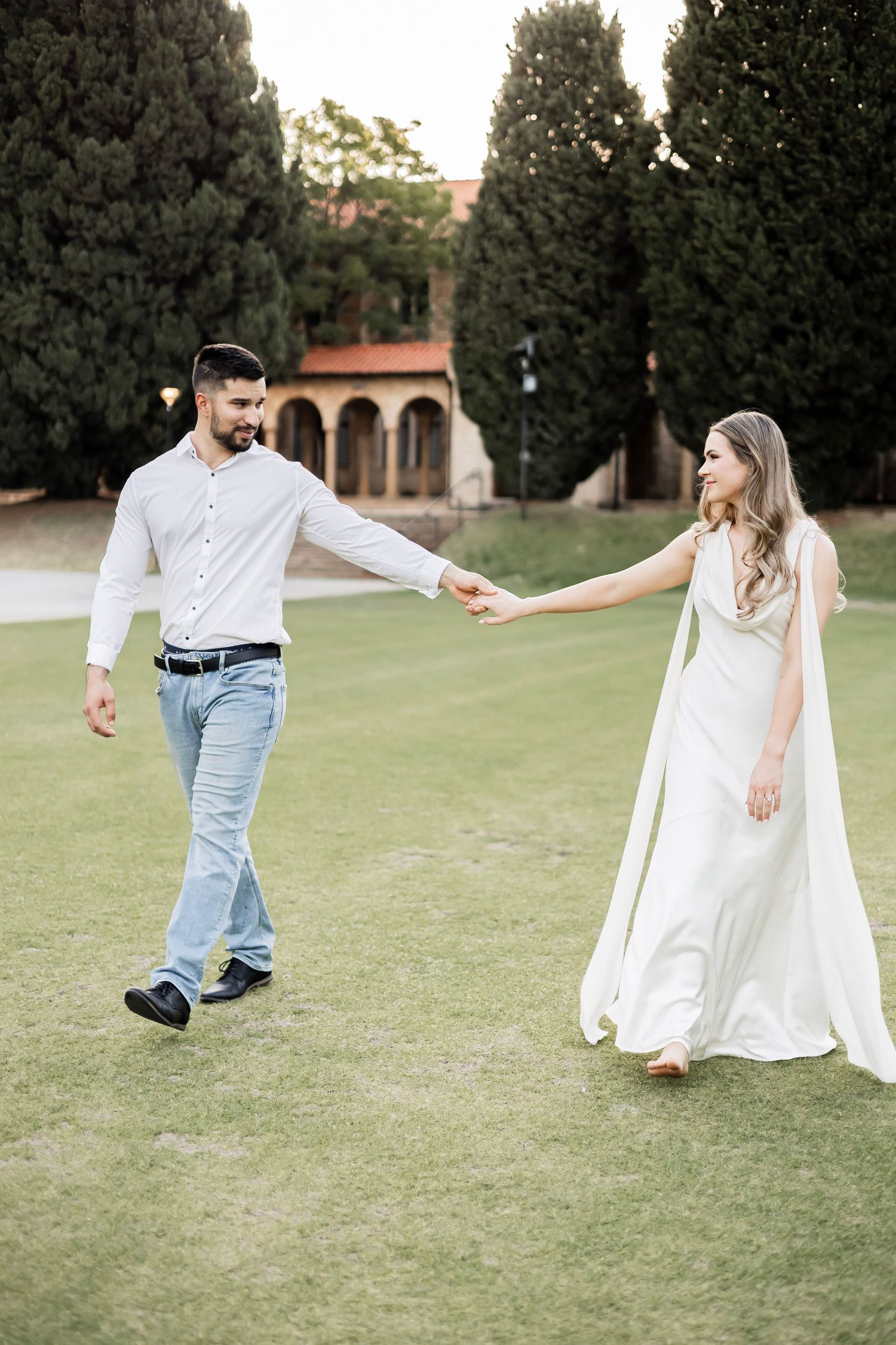 Couple holding hands and walking on green lawn with cypress trees and historic building in background.