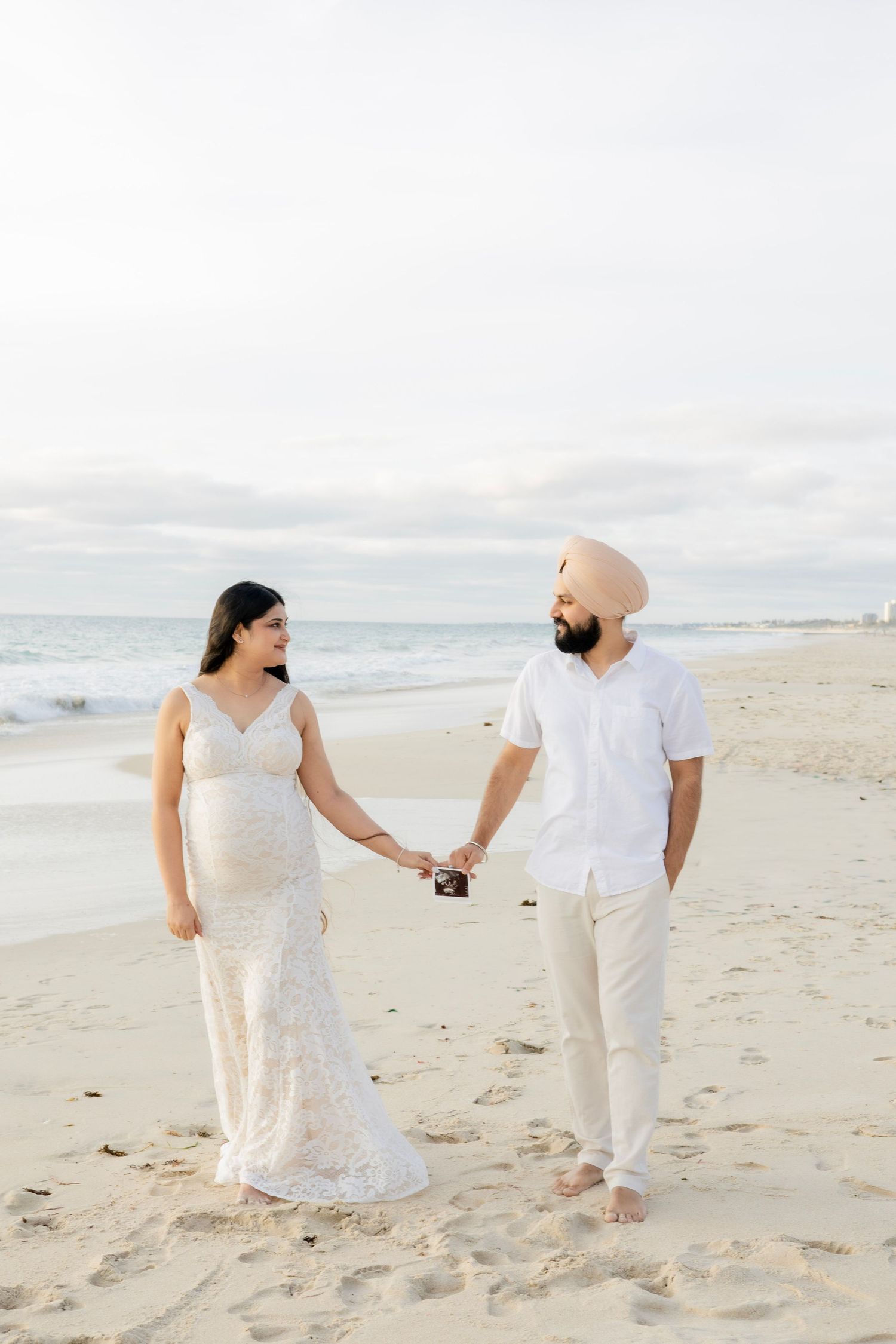 Expecting couple holding ultrasound photos while holding hands on a beach at sunset.