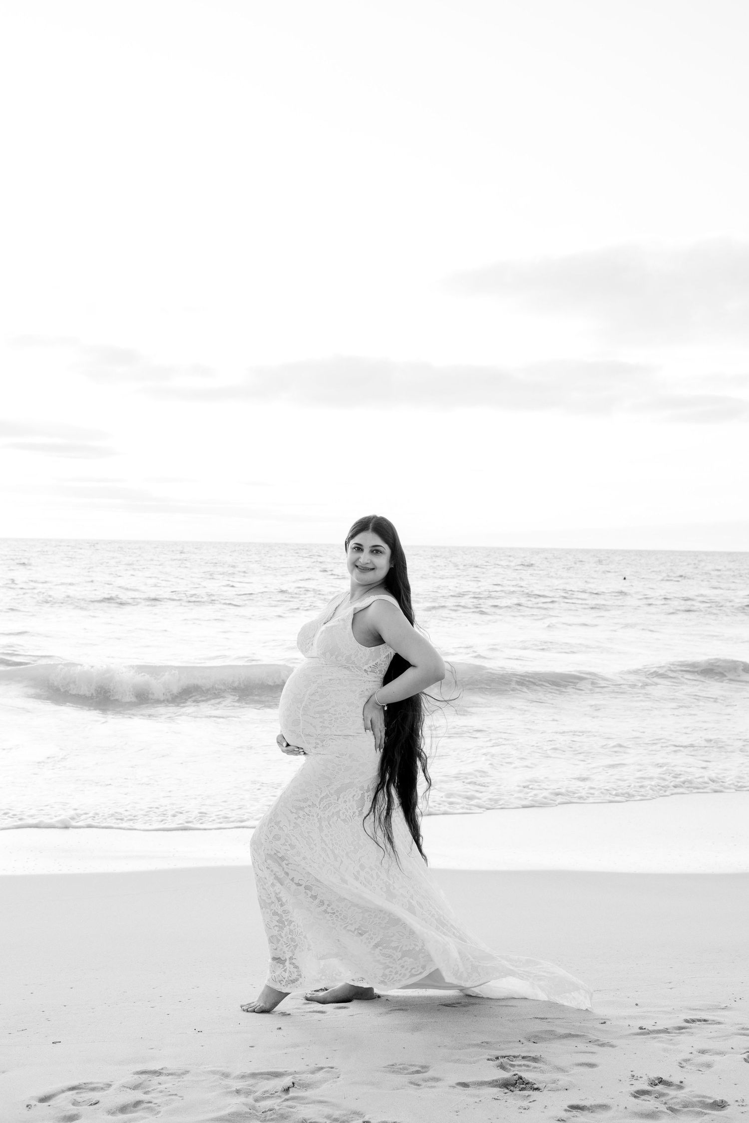 Black and white photo of pregnant woman in white lace dress posing on beach with ocean waves behind her.