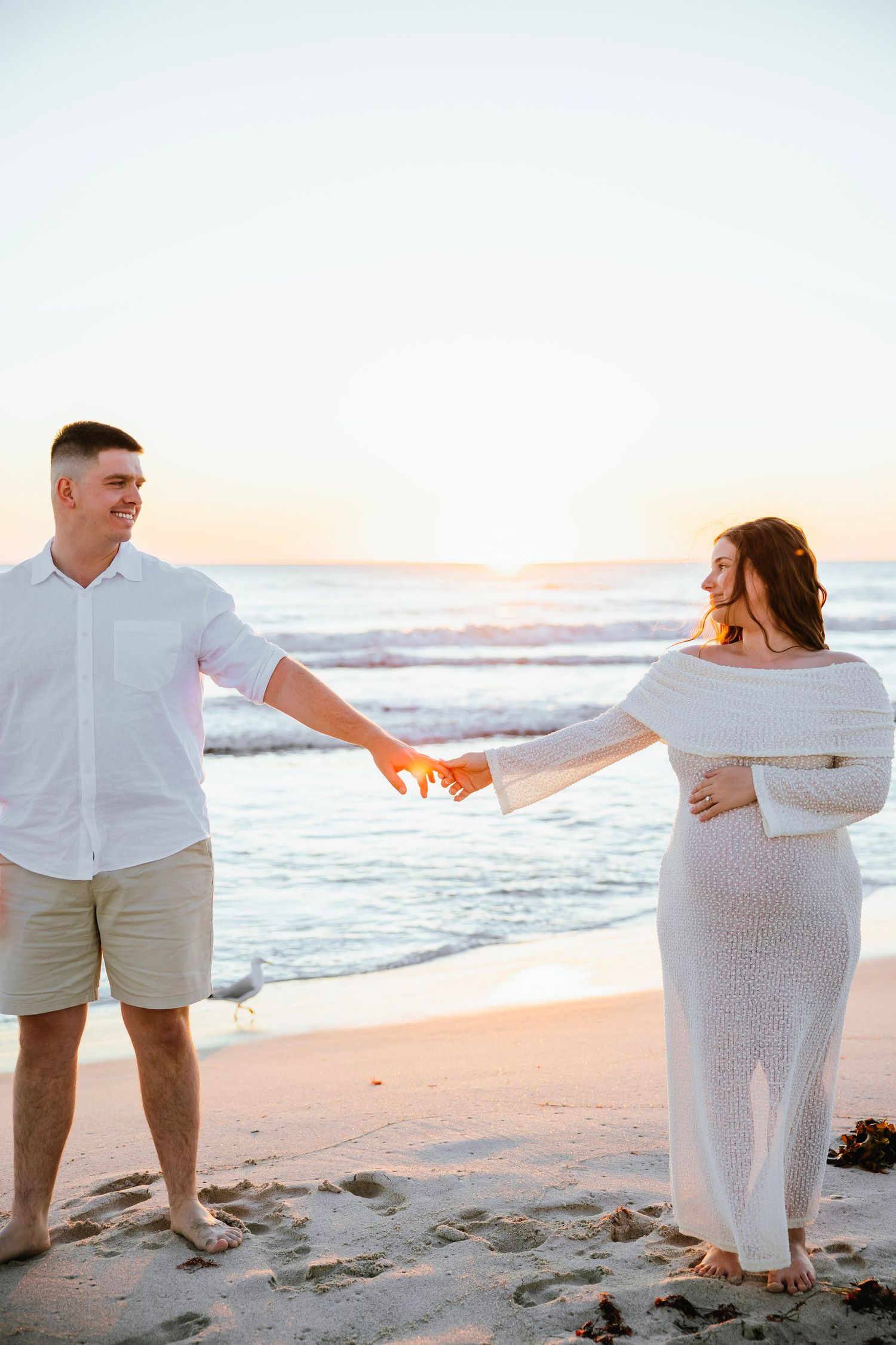 Pregnant couple holding hands on a sandy beach at sunset, woman in white dress, man in white shirt.