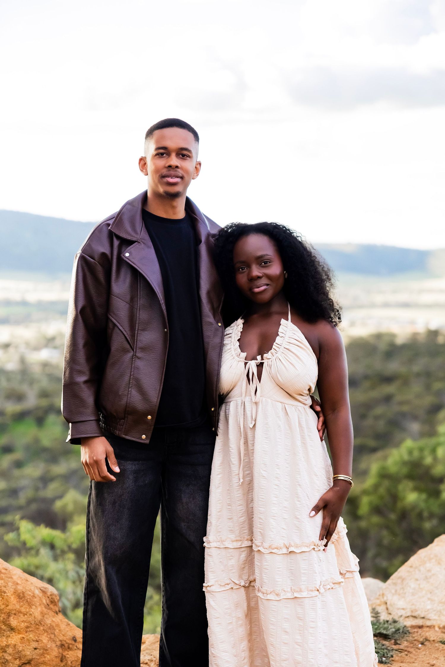 A young Black couple poses together on a rocky overlook, man in leather jacket and woman in white sundress, with scenic valley views.