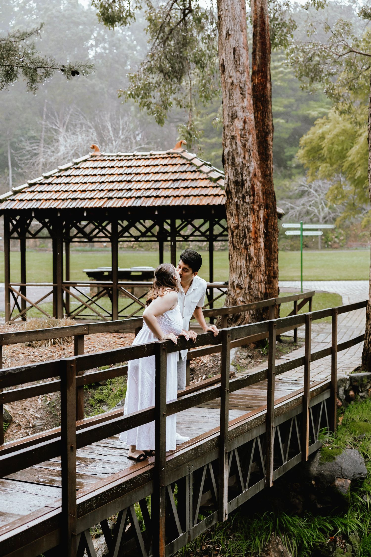 Couple embracing on a wooden bridge in a lush park with a gazebo in the background.