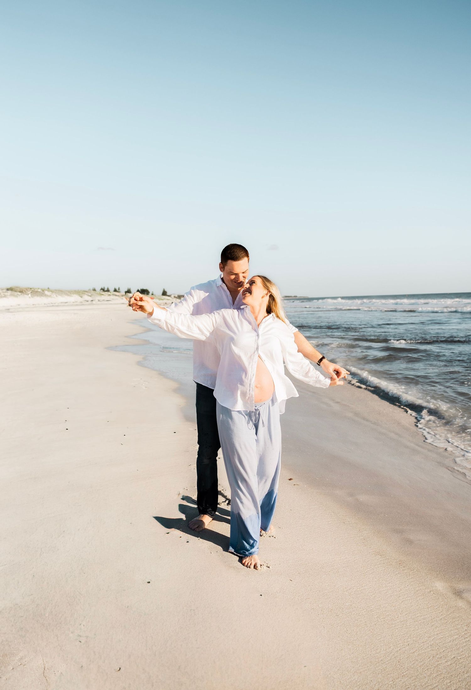 Happy pregnant couple dancing on sunny beach, woman in white shirt and blue pants, man in white shirt and jeans.