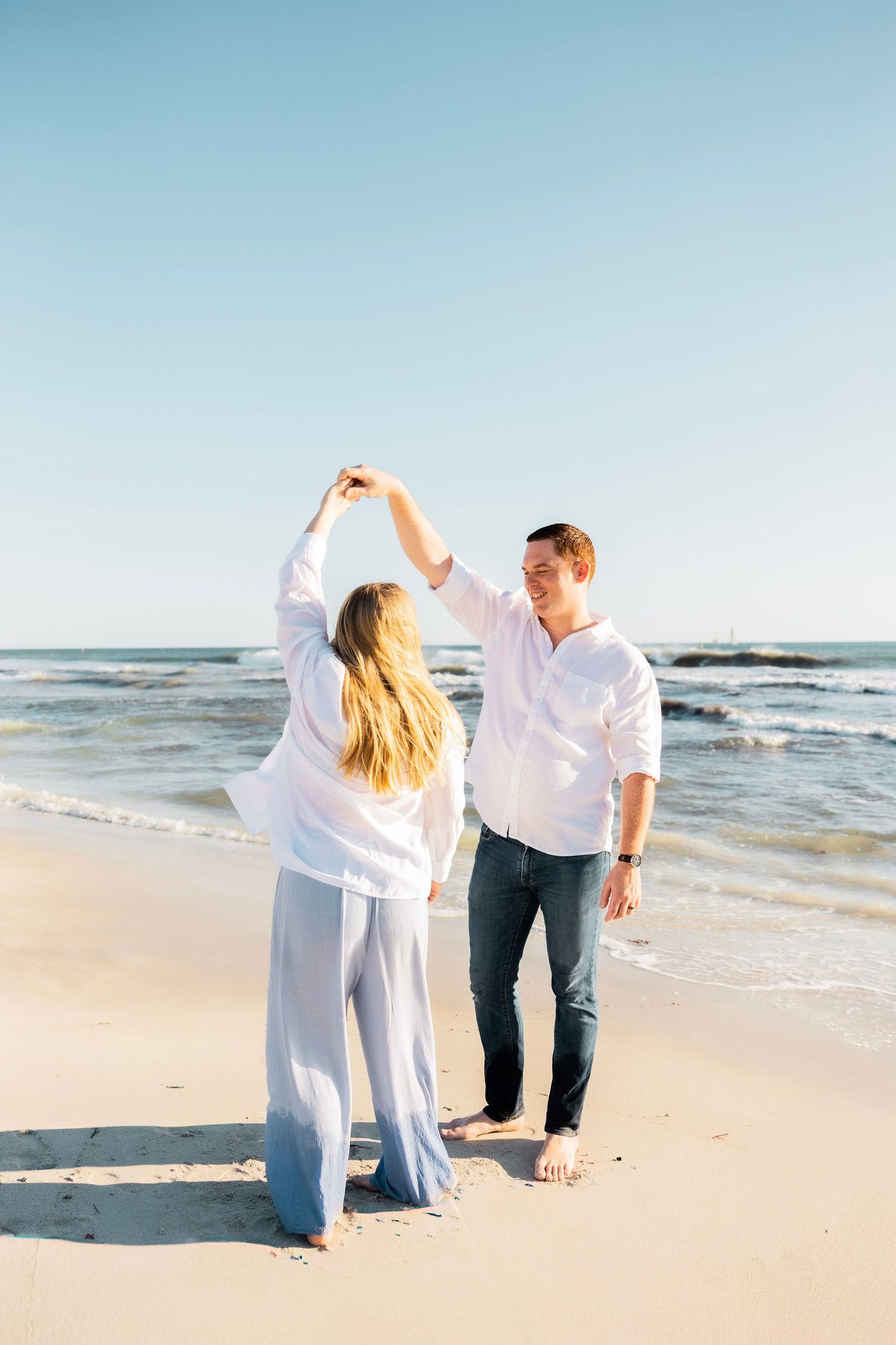 Couple dancing barefoot on sandy beach with ocean waves behind them.