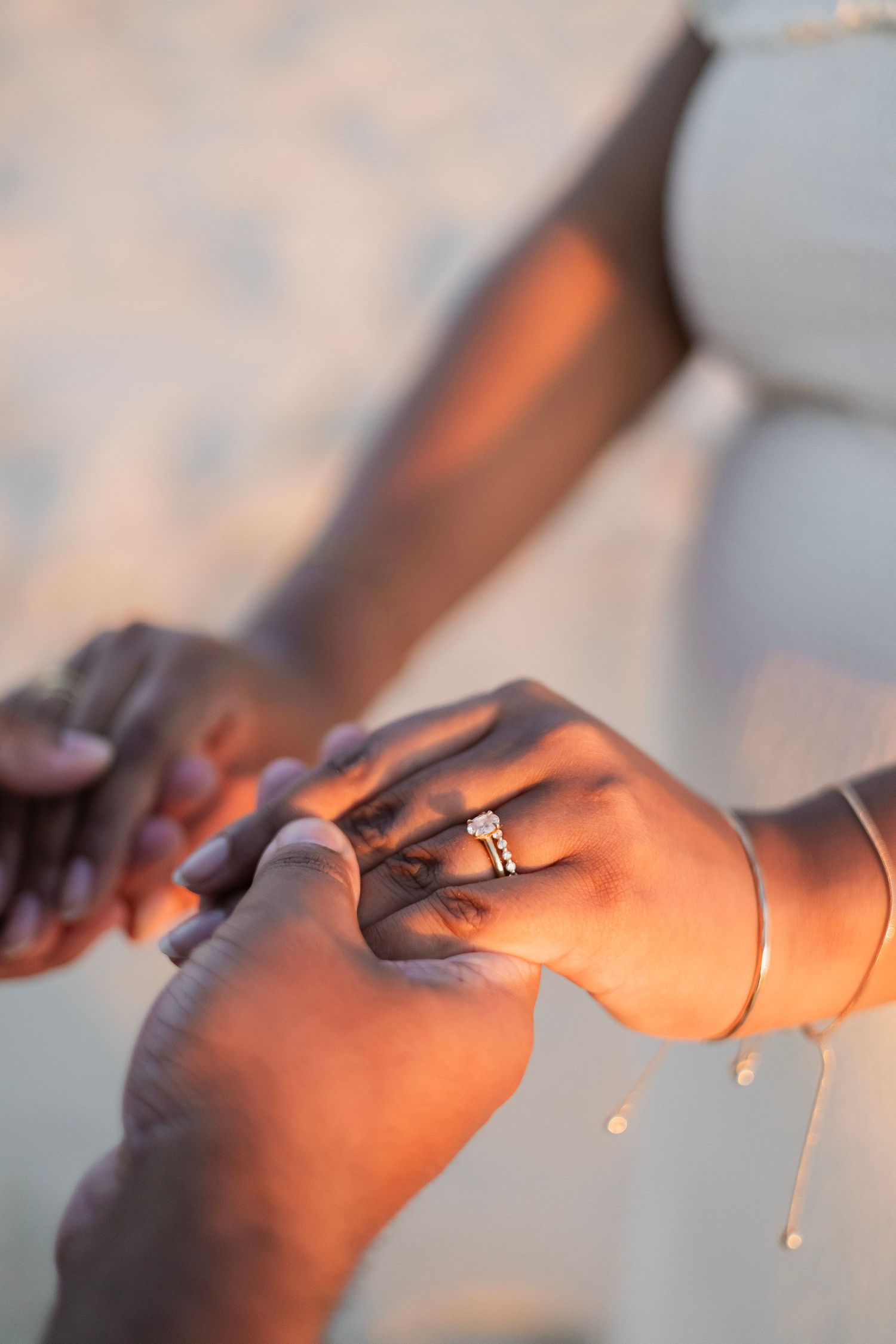 Close-up of hands holding on beach, woman wearing diamond engagement ring and gold bracelet in warm sunset light.