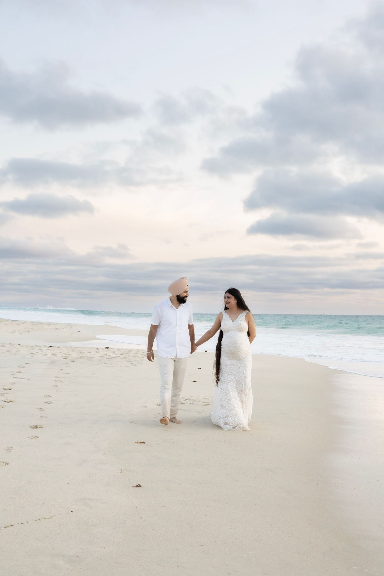 Expecting couple walking hand in hand along white sandy beach under dramatic cloudy sky.
