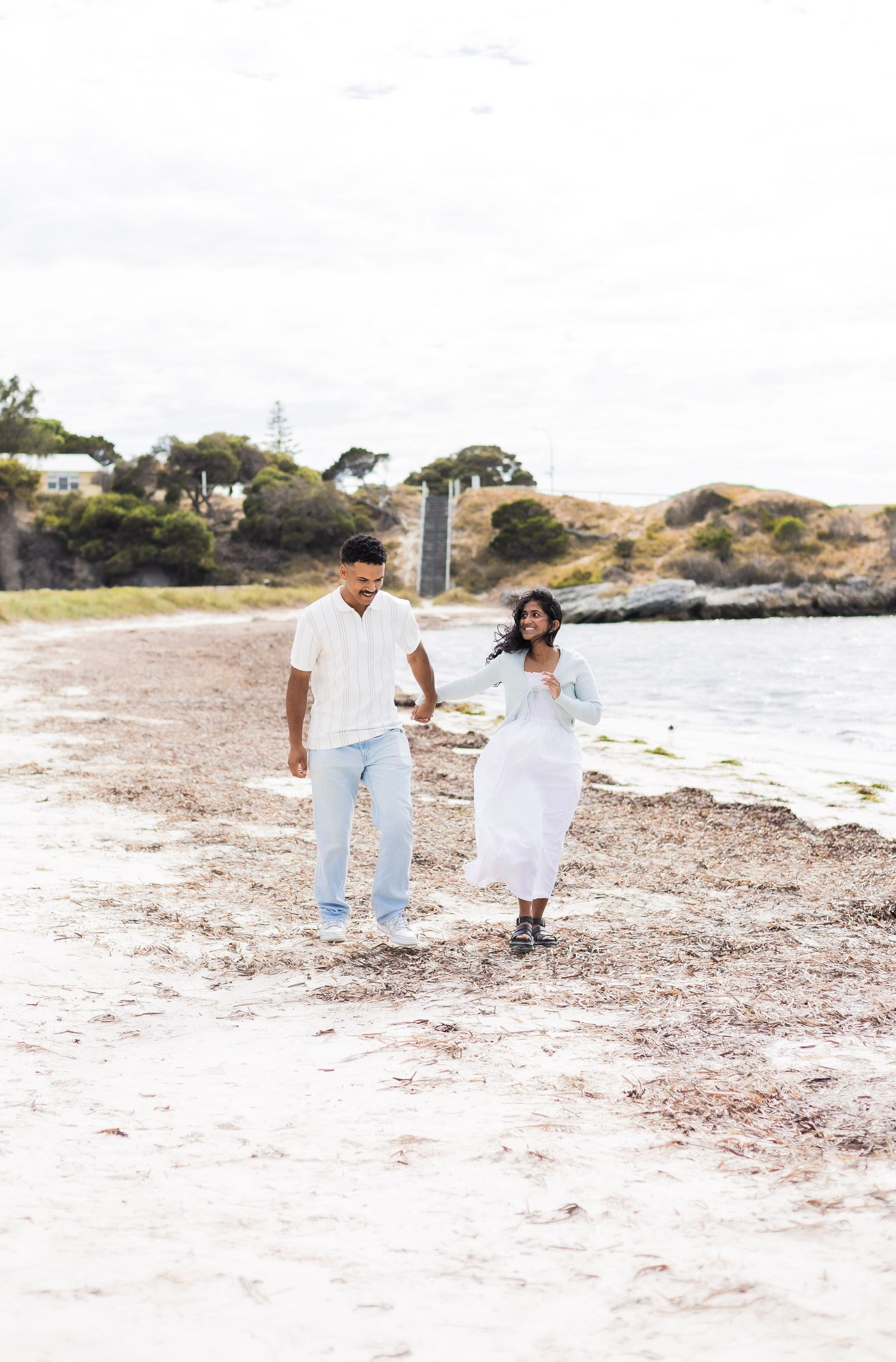 A couple walks hand in hand along a sandy beach shoreline with trees and water in the background.