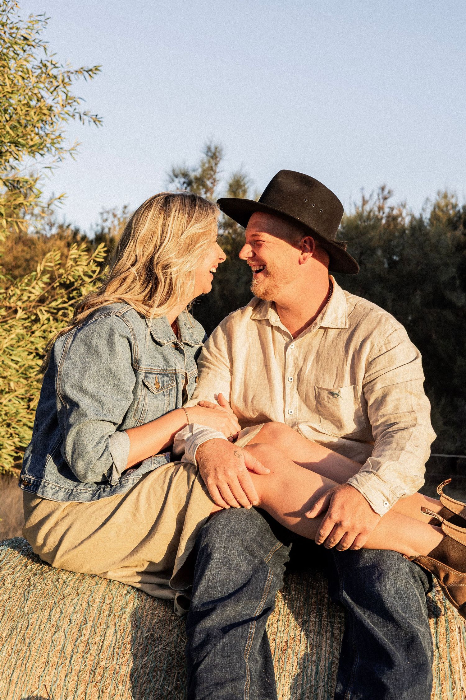 Smiling couple sitting together on a hay bale at golden hour surrounded by green trees.