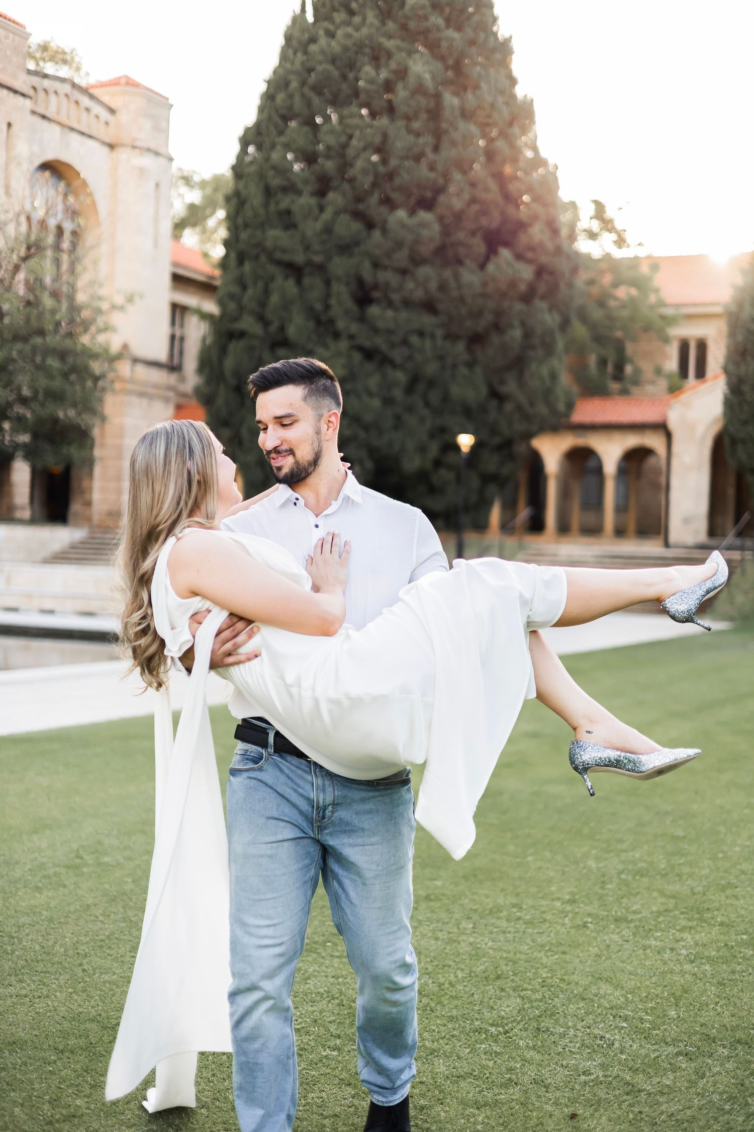 Man carrying woman in white dress in his arms while smiling on a green lawn near a building.