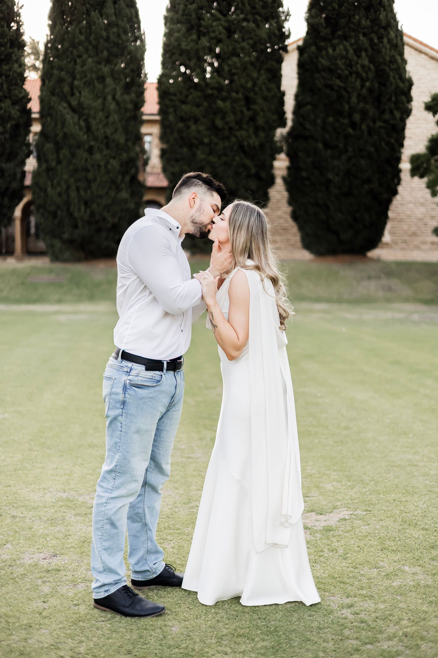 A couple shares a romantic kiss on a green lawn, with tall cypress trees and a building in the background.