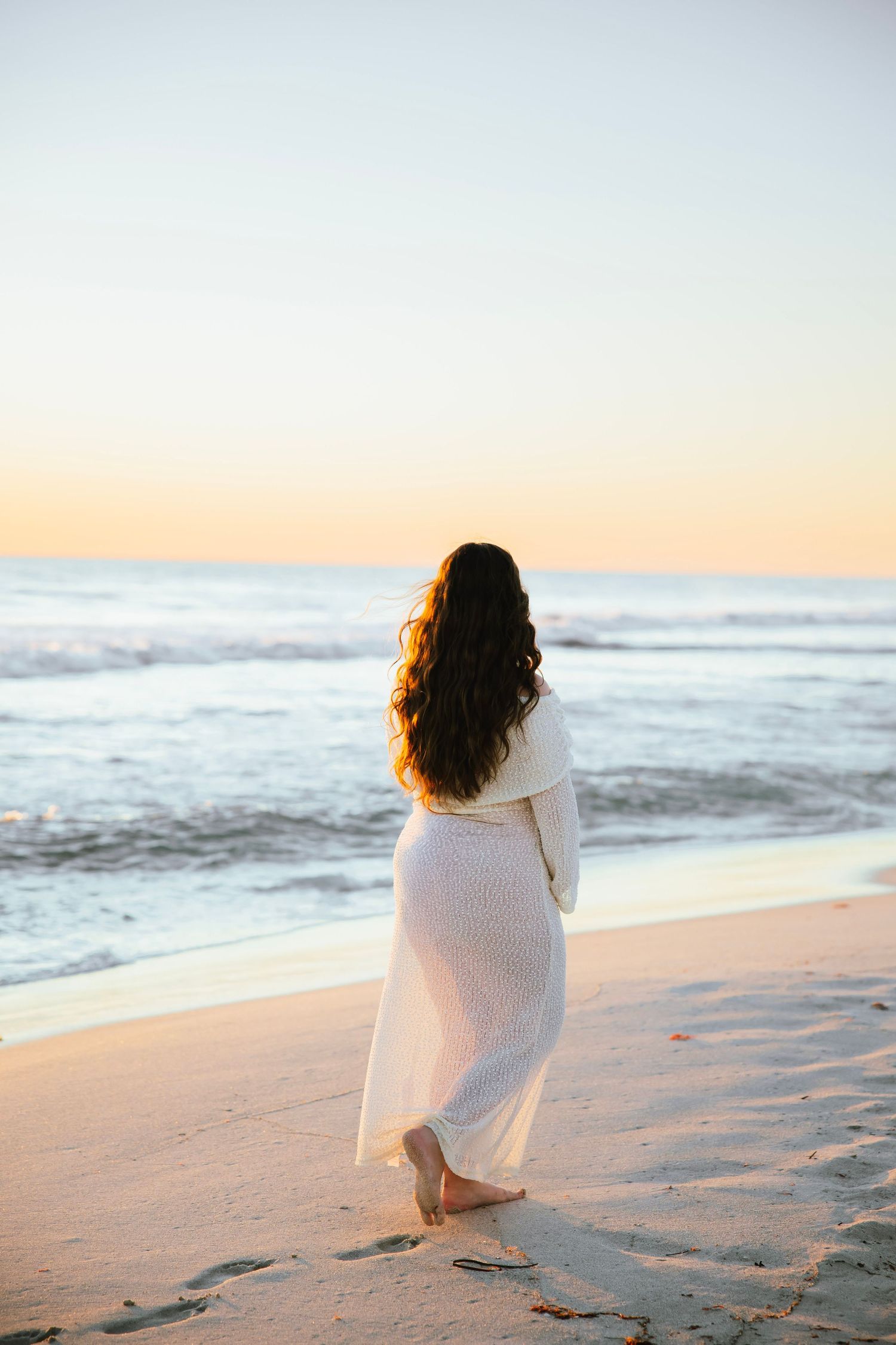 Pregnant woman in white knit dress standing barefoot on beach at sunset, facing ocean waves.