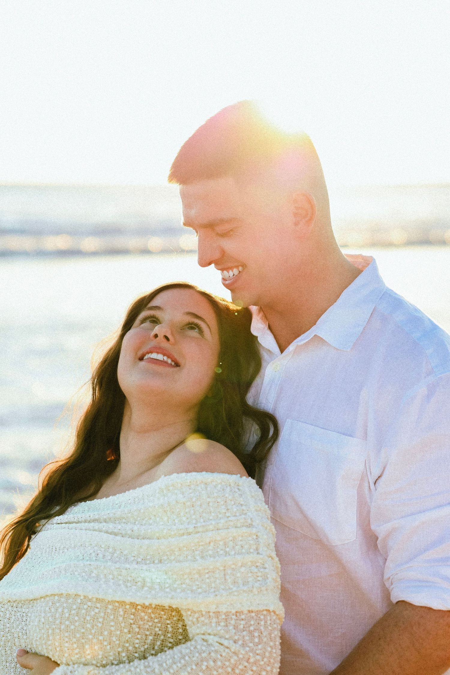 Happy couple sharing a romantic moment on a sunny beach, smiling at each other in white outfits with ocean backdrop.