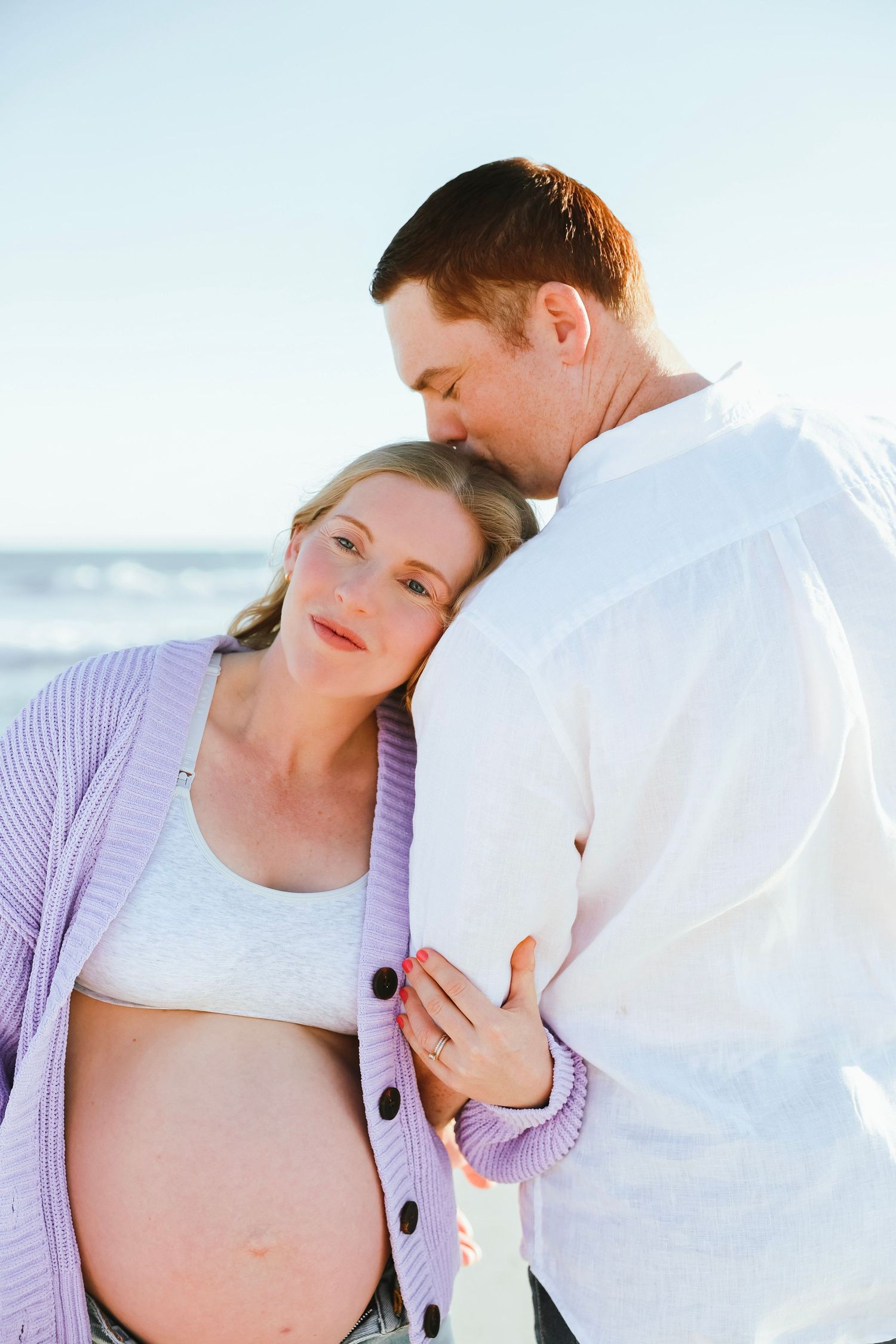 Pregnant woman in lavender cardigan leaning on partner's shoulder at beach, man kissing her head, sunny day.