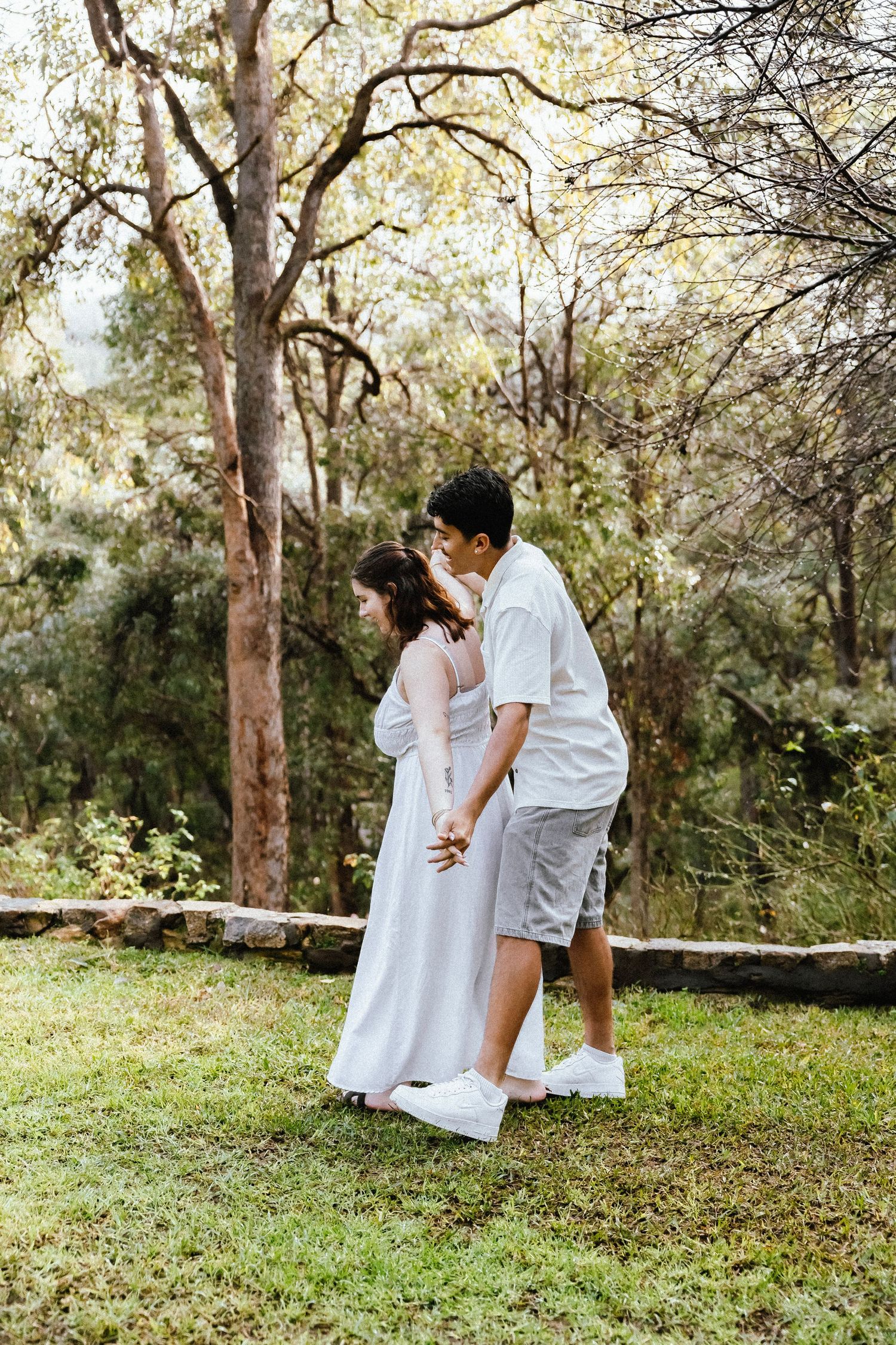 Couple walking hand in hand through a lush green forest with tall trees surrounding them.