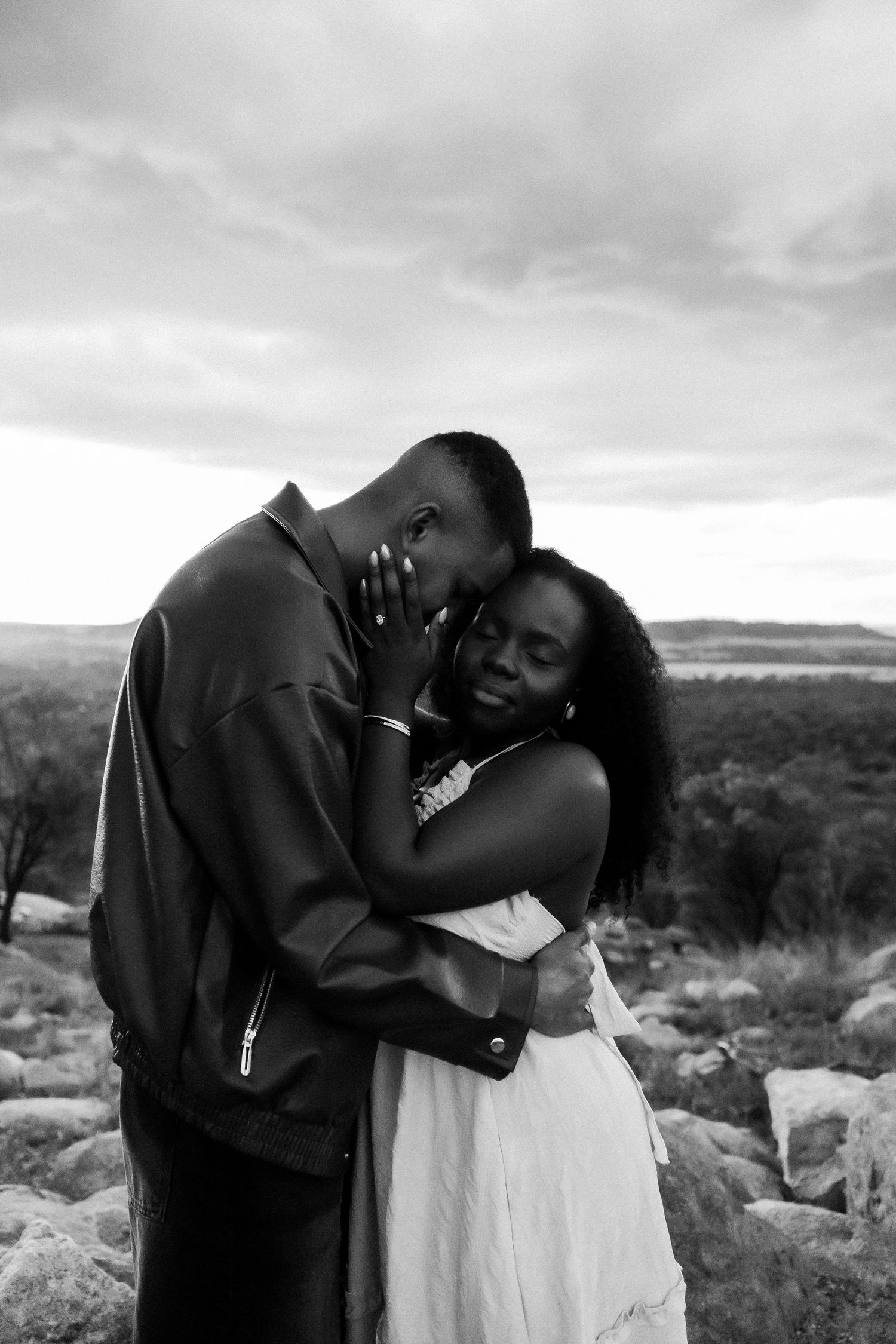 Black and white photo of a couple embracing tenderly outdoors on rocky terrain with a cloudy sky backdrop.