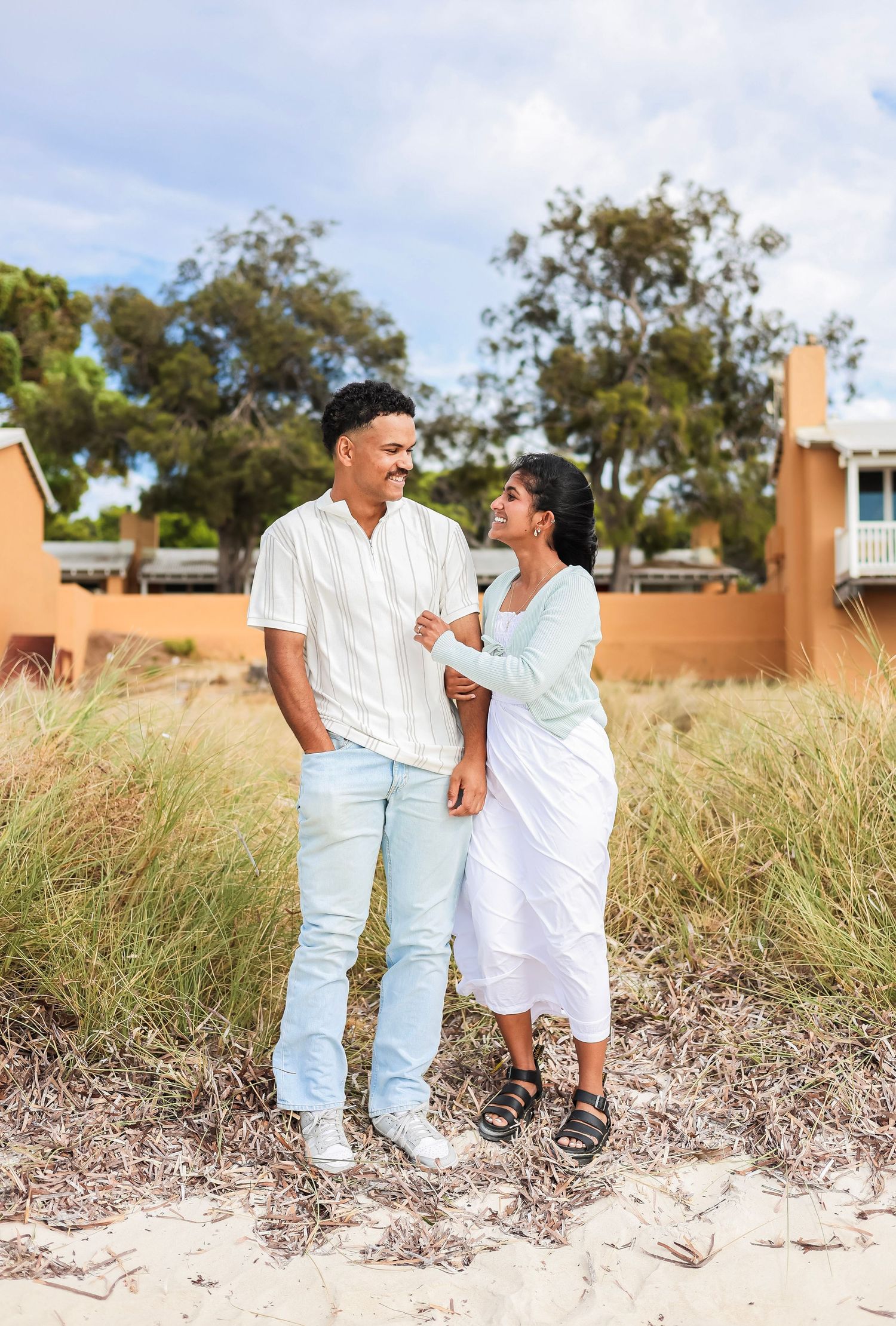 Happy couple standing on beach sand dunes, smiling at each other surrounded by coastal greenery.