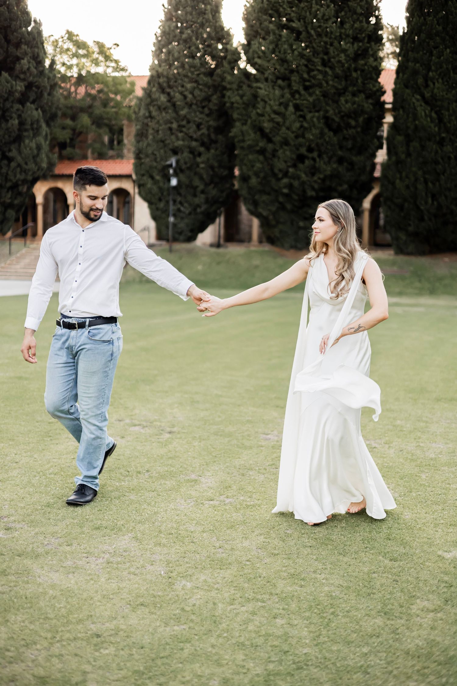 Couple holding hands and strolling on green lawn with tall cypress trees and historic building backdrop.