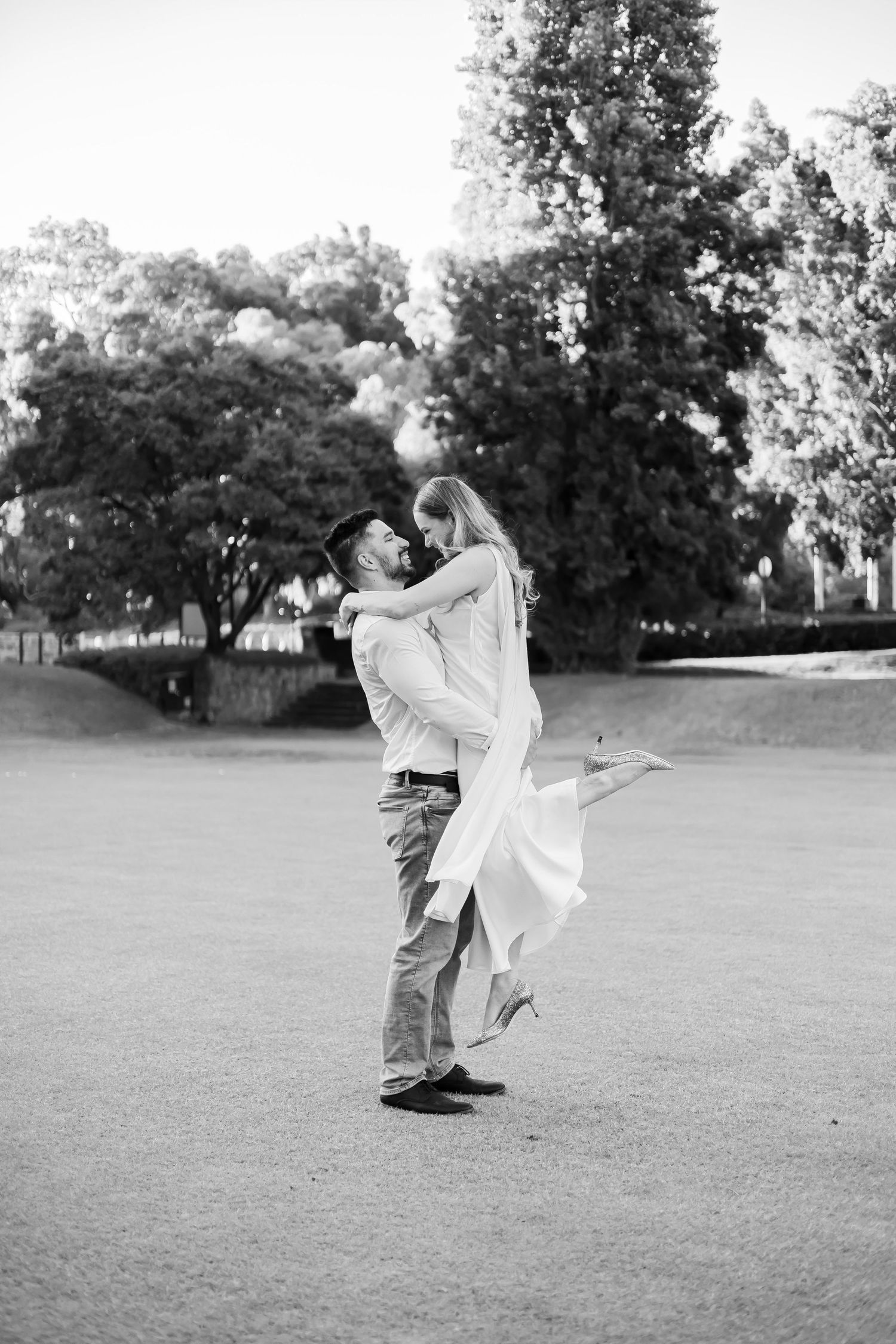 Man lifting woman in white dress outdoors on grass with trees in black and white engagement photo.
