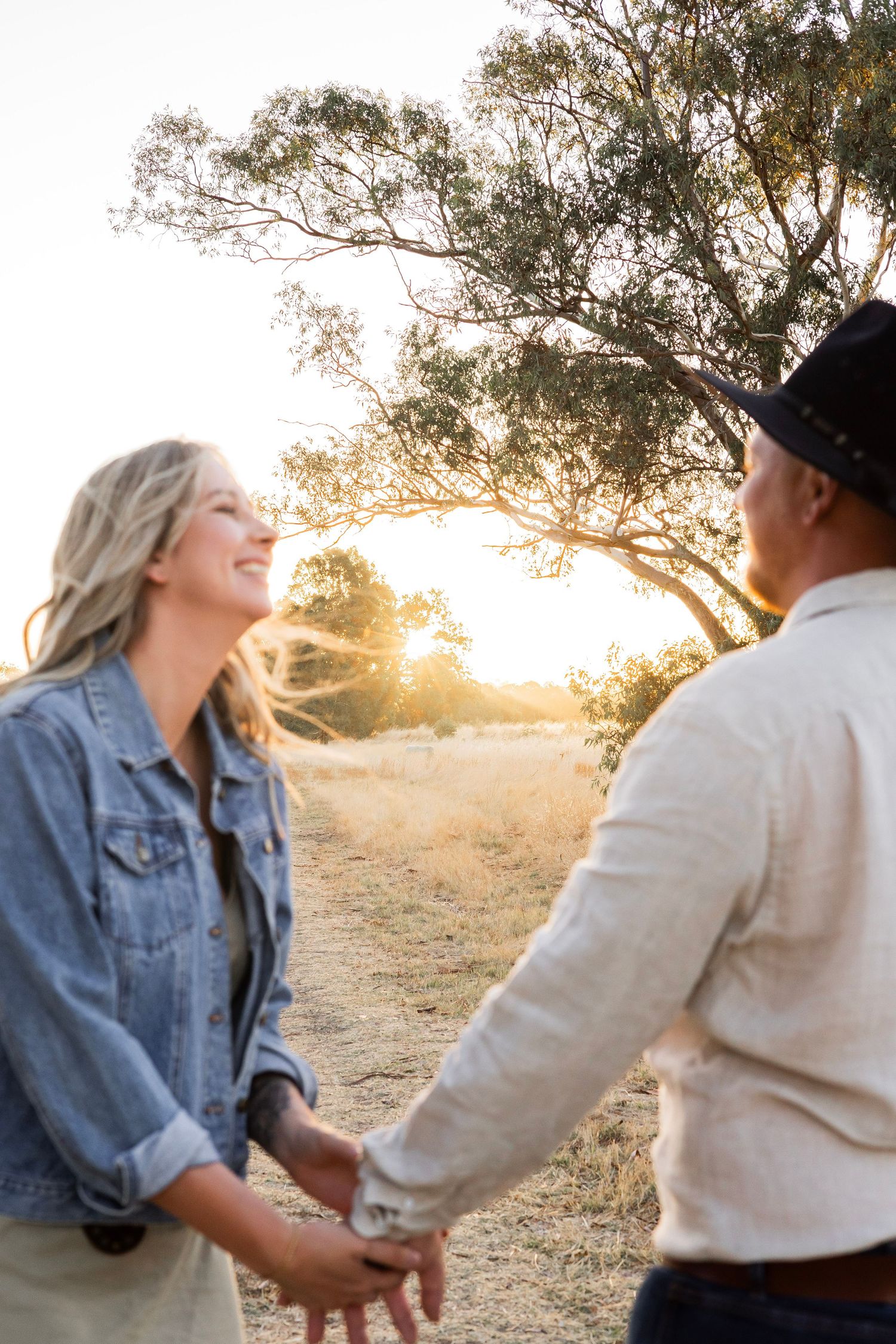 Couple holding hands in a golden field at sunset, woman laughing joyfully in a denim jacket.
