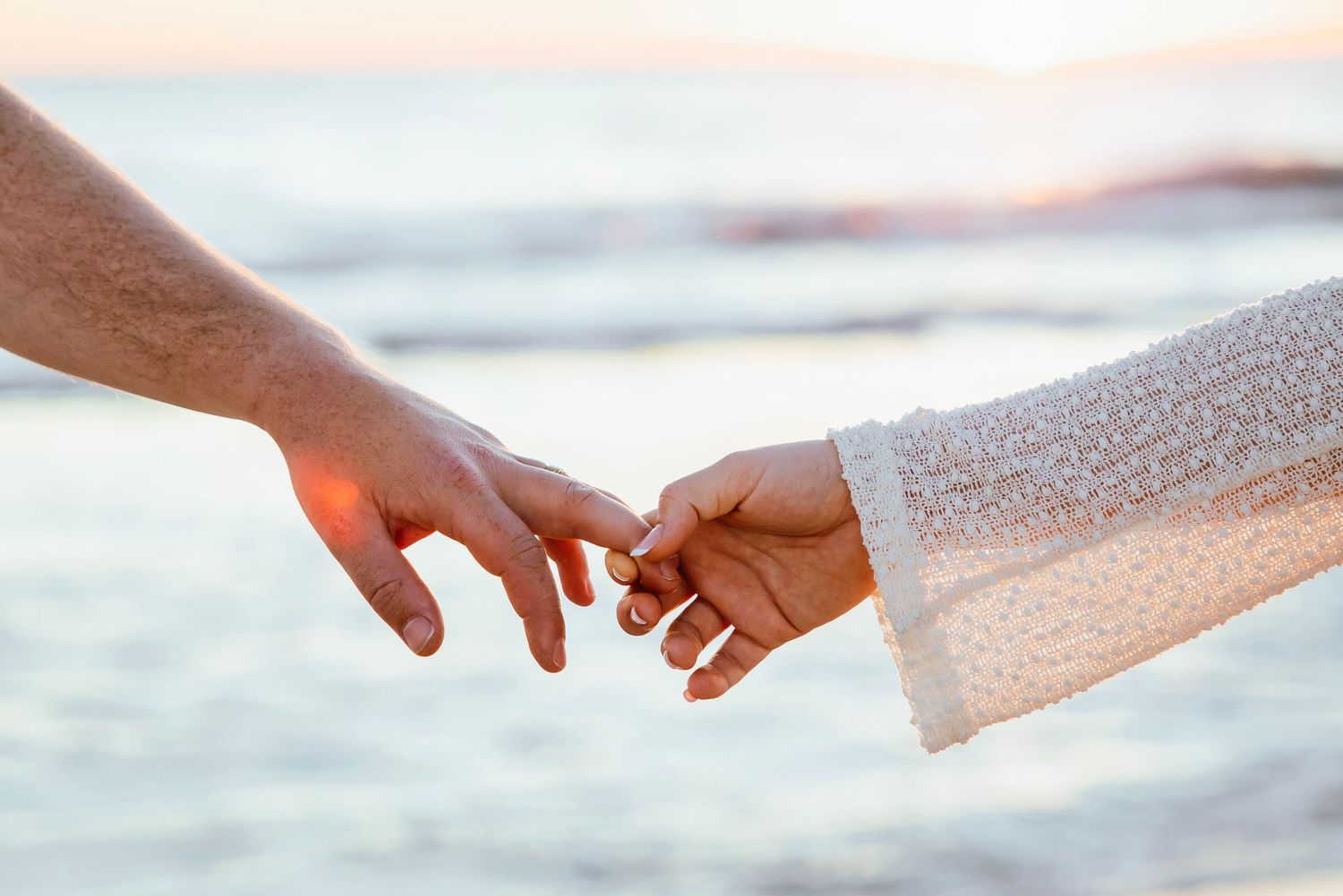 Couple holding hands tenderly at beach sunset, woman wearing white lace sleeve with ring visible.