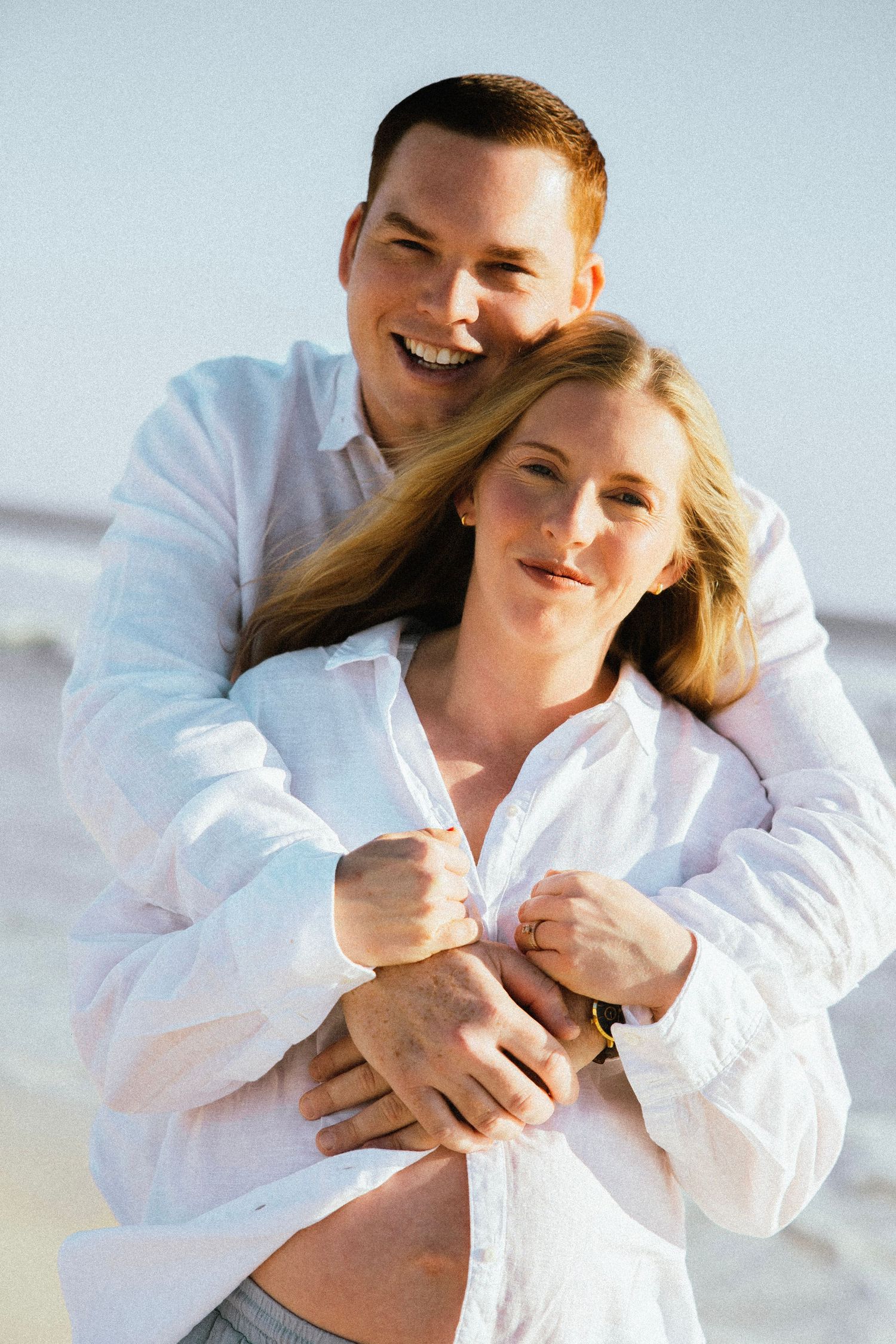 A smiling couple in white shirts embraces on a sunny beach, the man wrapping his arms around the pregnant woman.