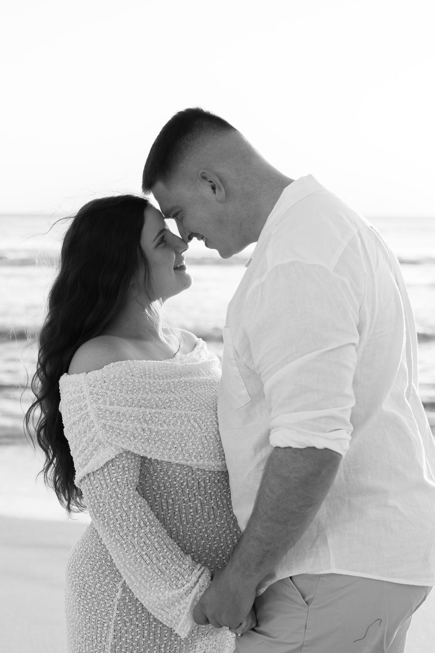 Black and white photo of a couple touching foreheads at the beach, woman in off-shoulder dress with baby bump.